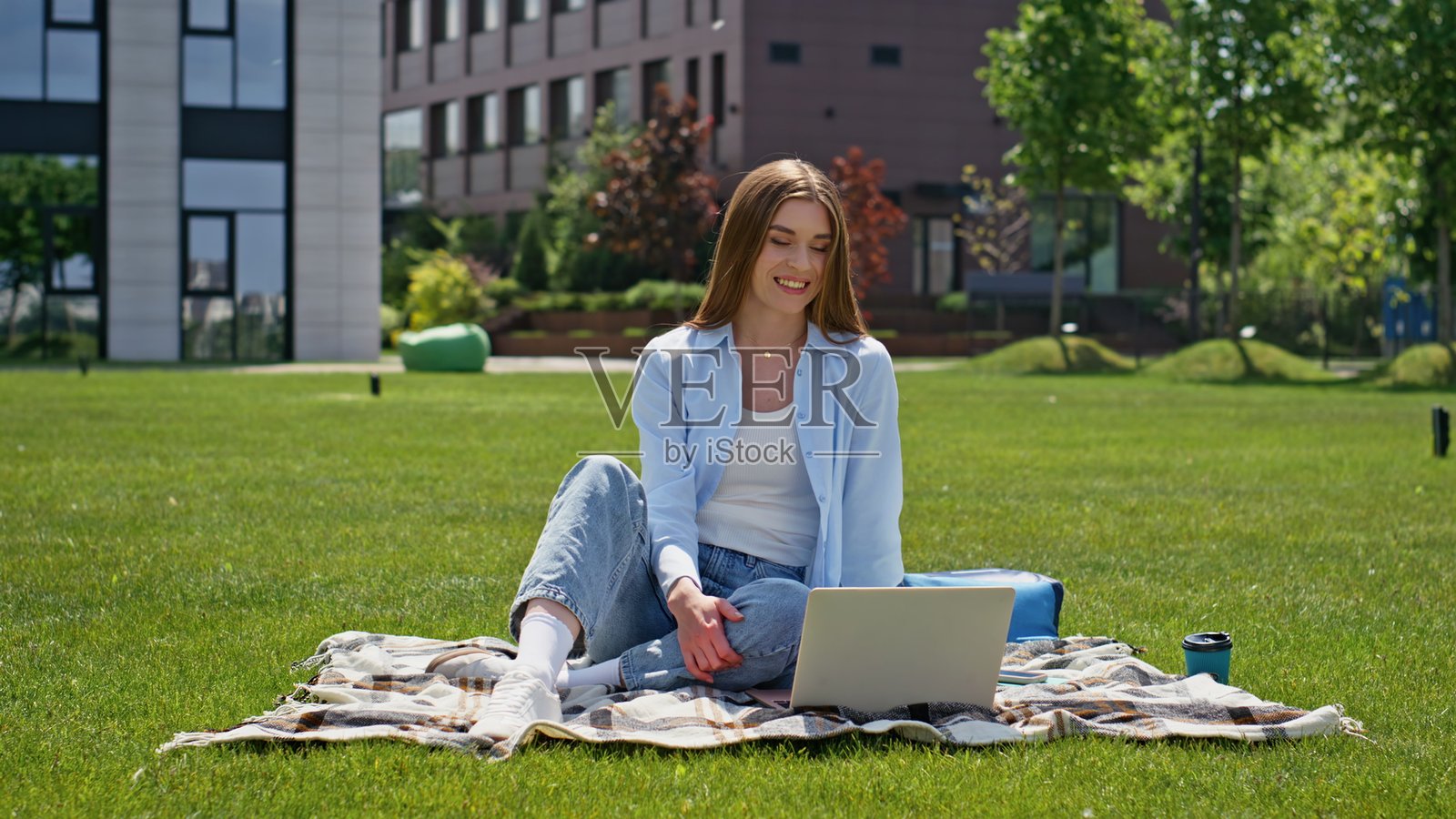 微笑的女孩坐在绿色草坪上用笔记本电脑视频通话。快乐的女人照片摄影图片