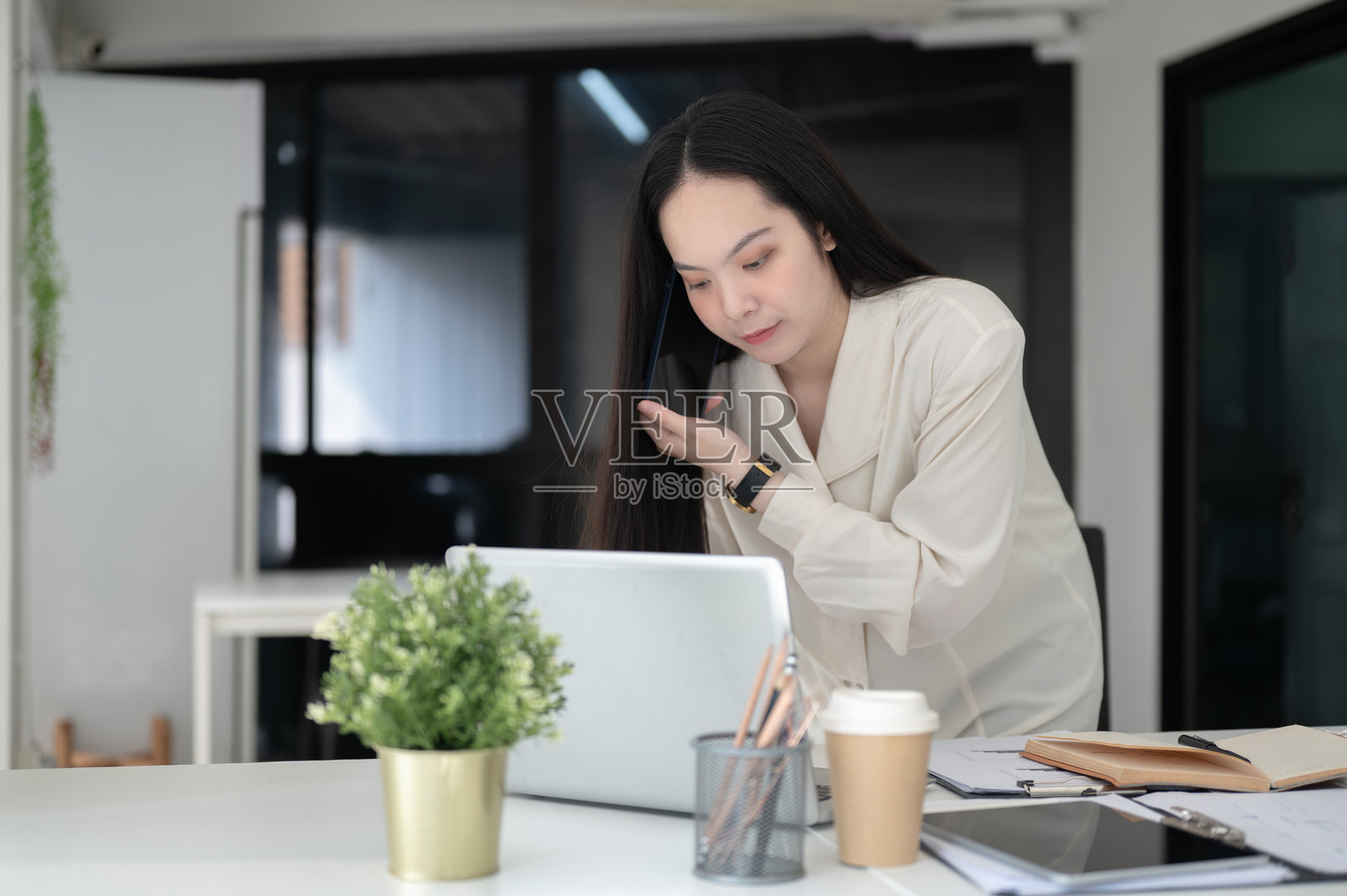 女商人或女性办公室工作人员一边用智能手机通话，一边倾身查看桌上的笔记本电脑。照片摄影图片