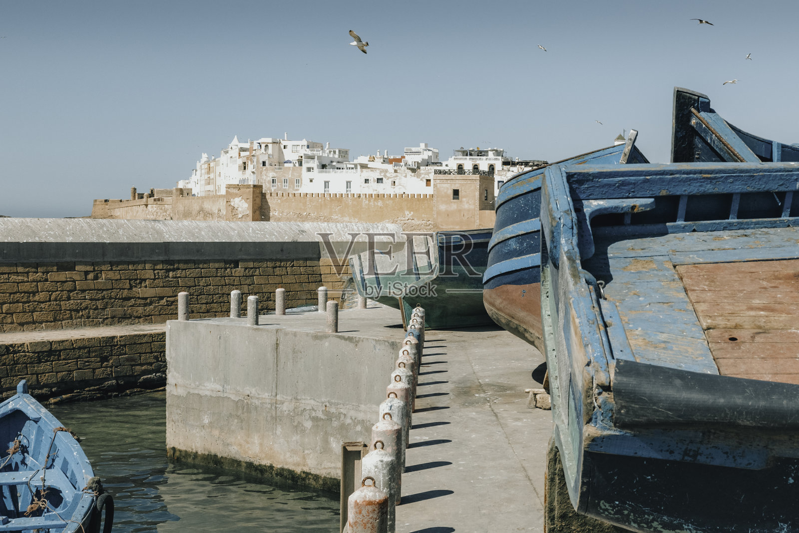 摩洛哥埃萨乌伊拉的港口停靠着传统的蓝色渔船，背景是历史悠久的城墙堡垒，阳光明媚，展现了这个海滨小镇的迷人港口景色。照片摄影图片
