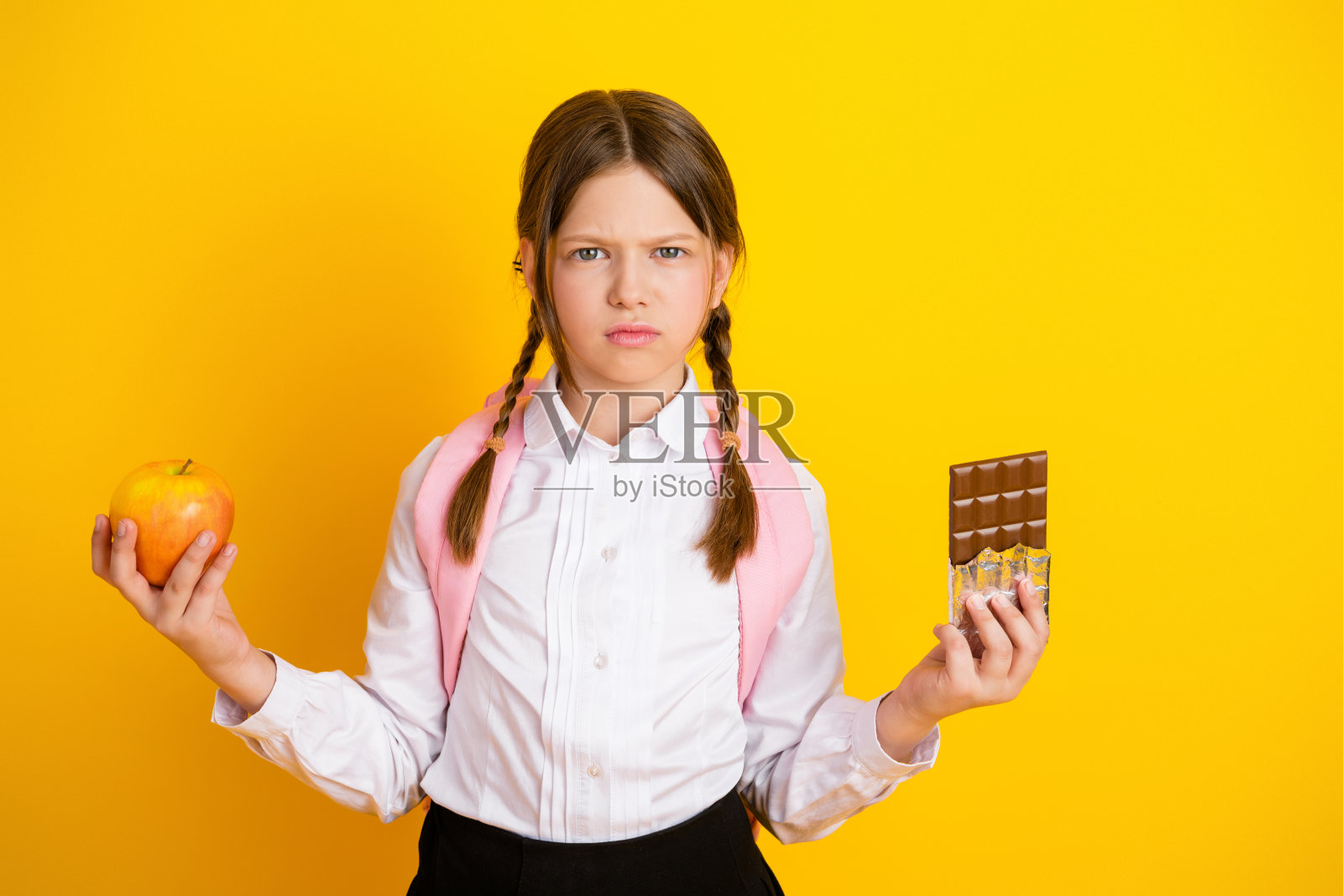 年轻的女学生手持苹果和巧克力，展示选择，背景为明亮的黄色，适合家庭或教育环境。照片摄影图片
