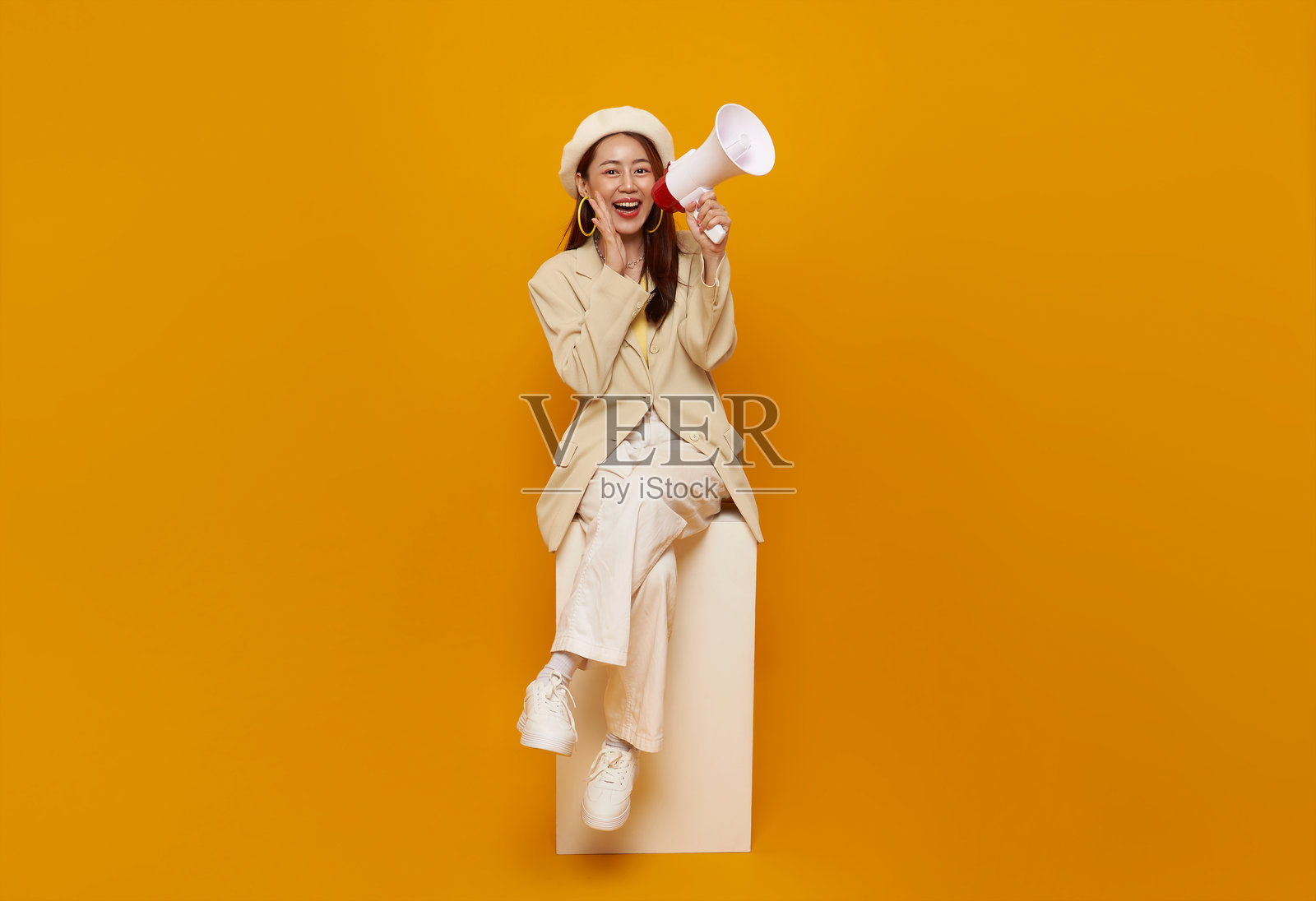 年轻的亚洲女商人坐在白色箱子上，手持扩音器在黄色背景下大声喊叫。演讲和宣布的概念。照片摄影图片