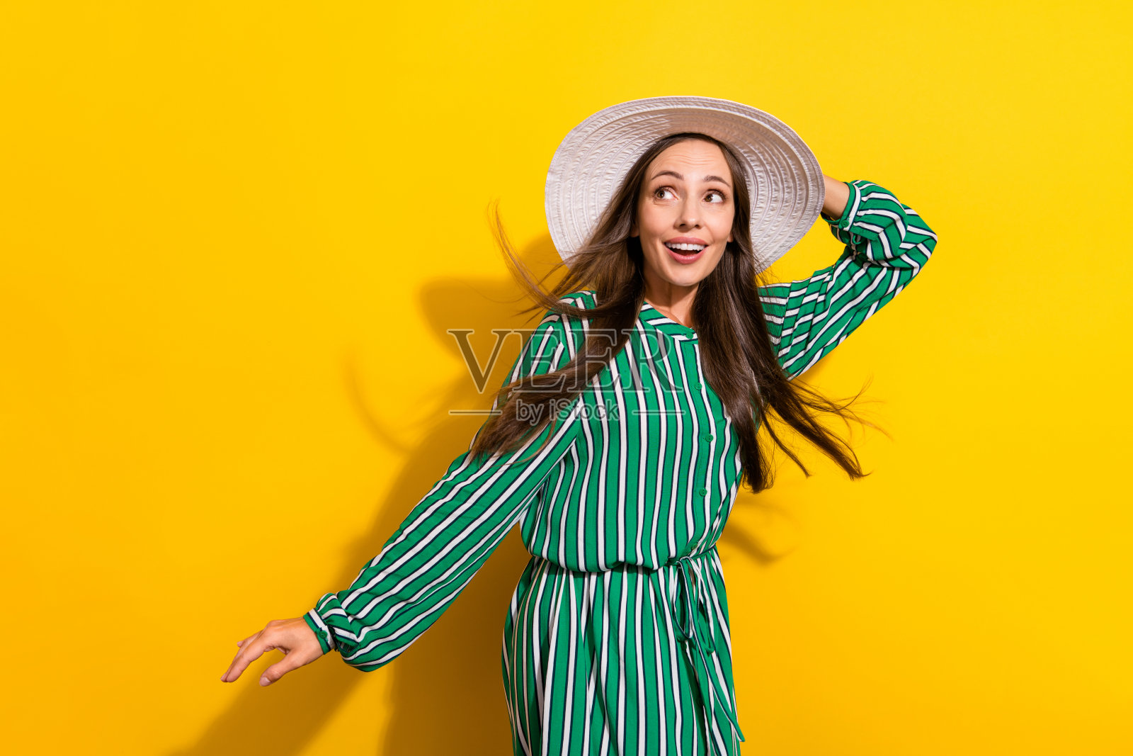 一位成熟的深棕色头发女士穿着绿色连衣裙，佩戴帽子，惊讶地看着空旷的空间，背景为黄色。照片摄影图片