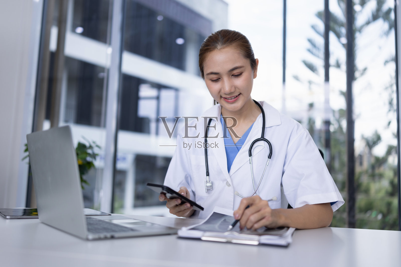 快乐微笑的亚洲女性医生在医院的桌子上与患者的X光片和笔记本电脑一起工作。照片摄影图片