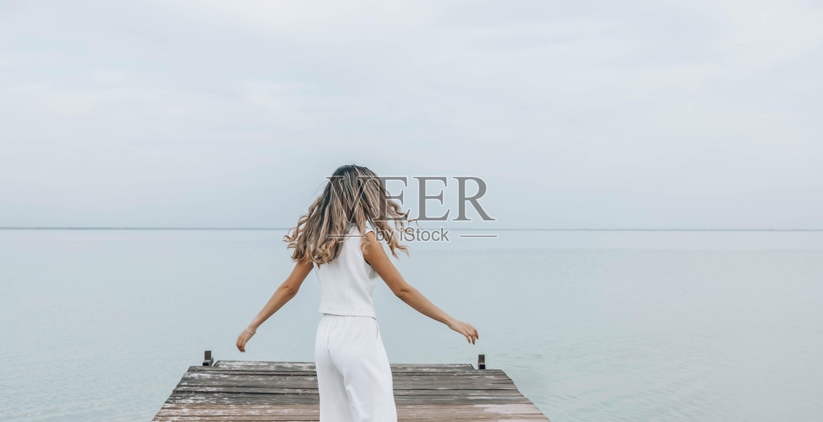 一个无法辨认的女人在木栈道上跳舞，长发随风飘扬，轻松旋转，苍白的天空，四周被宁静的水域环绕，面带快乐的表情，展现自由与健康，运动模糊。照片摄影图片