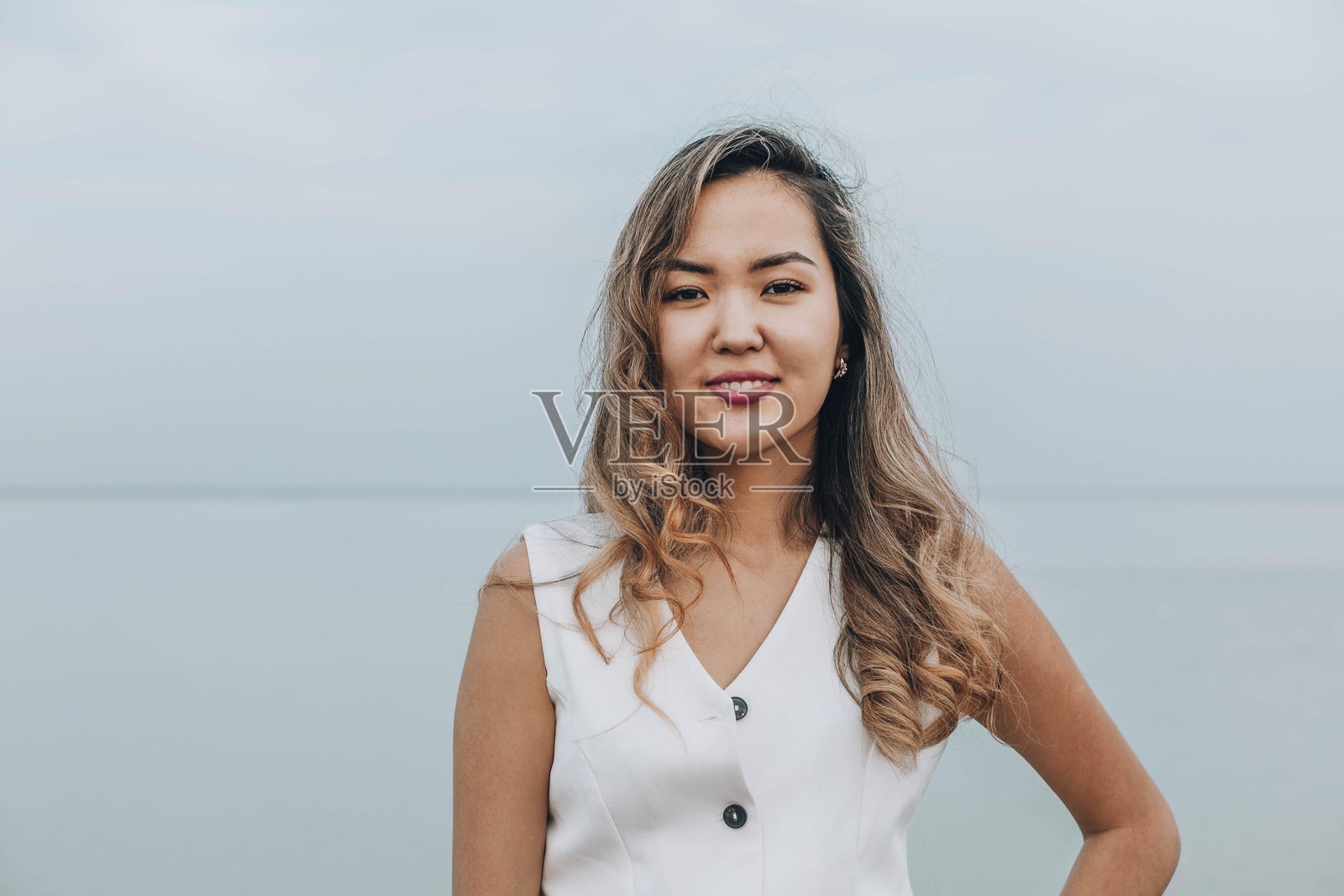 亚洲女性微笑着看向镜头，坐在宁静的湖边，穿着白色服装，展现出自信和愉悦的情绪，体现健康、护肤和个人发展的概念。照片摄影图片