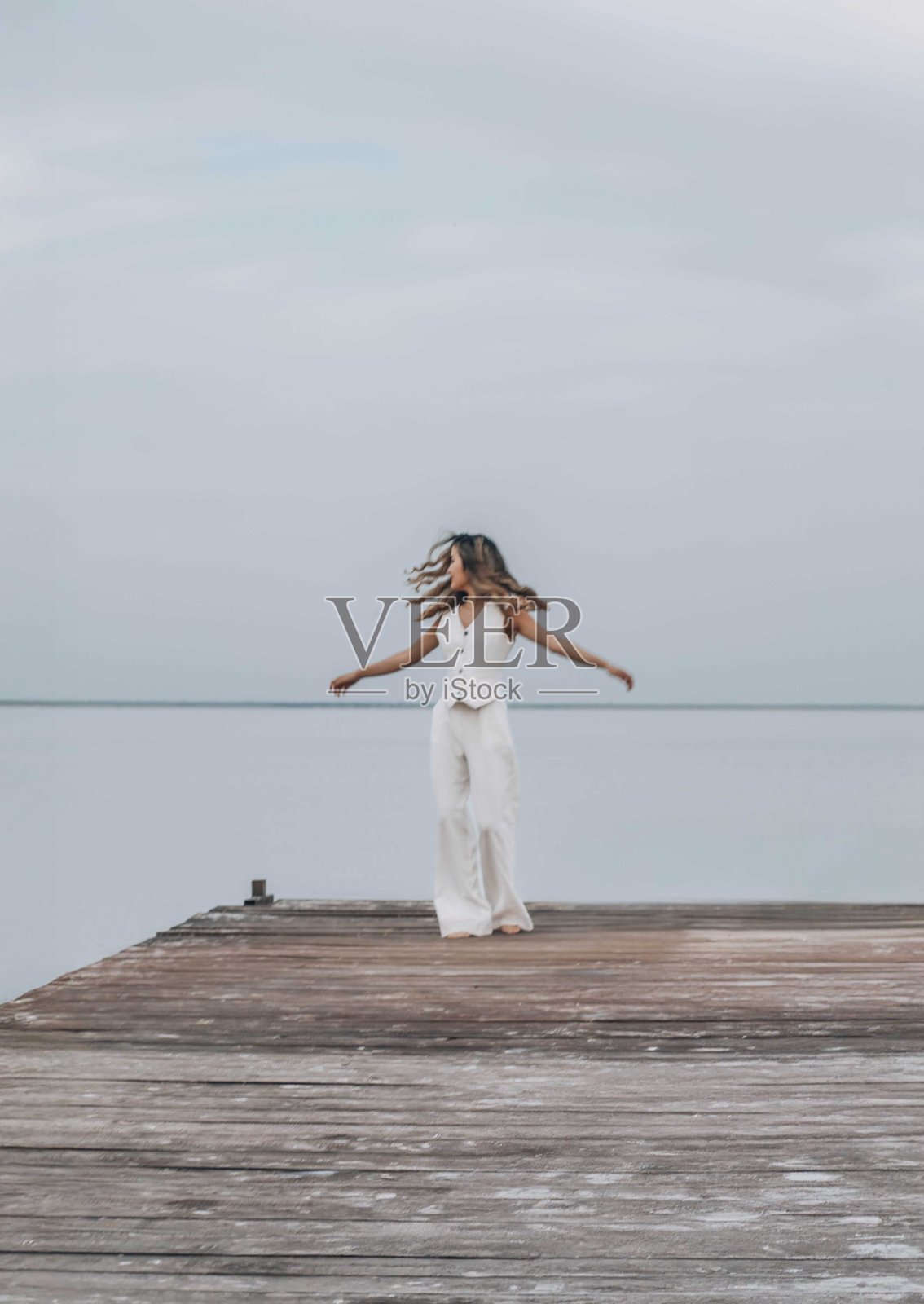 赤脚在木栈道上跳舞的女人，长发随风飘扬，她在淡蓝色的天空下自由旋转，周围是宁静的水面，脸上洋溢着快乐的表情，象征着自由与健康，动态模糊。照片摄影图片