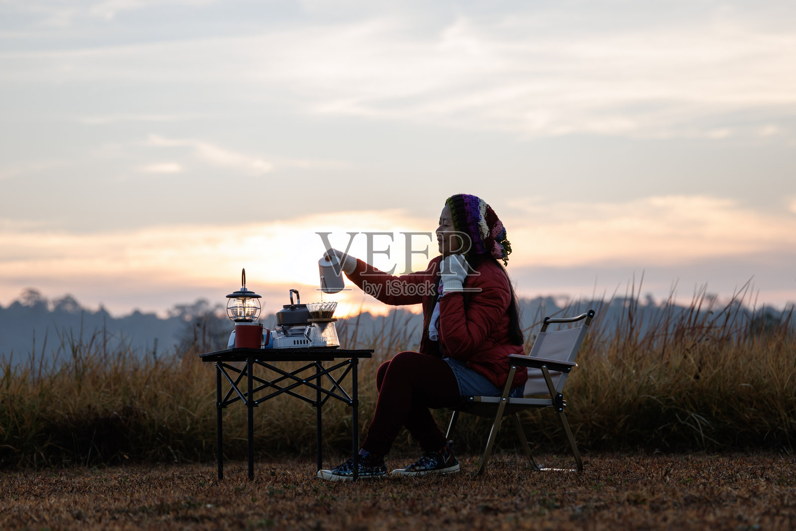 女人坐在户外，周围有露营装备，包括桌子、椅子和一盏蒸汽灯。她在宁静自然的环境中享受早晨的咖啡。照片摄影图片