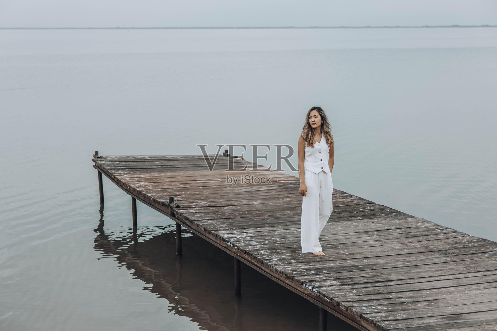 身穿白色服装的亚洲女性站在木栈道上，宁静的湖面，平和的阴天，从背后看，流露出沉思的情绪，正念，旅行静修，身心健康照片摄影图片