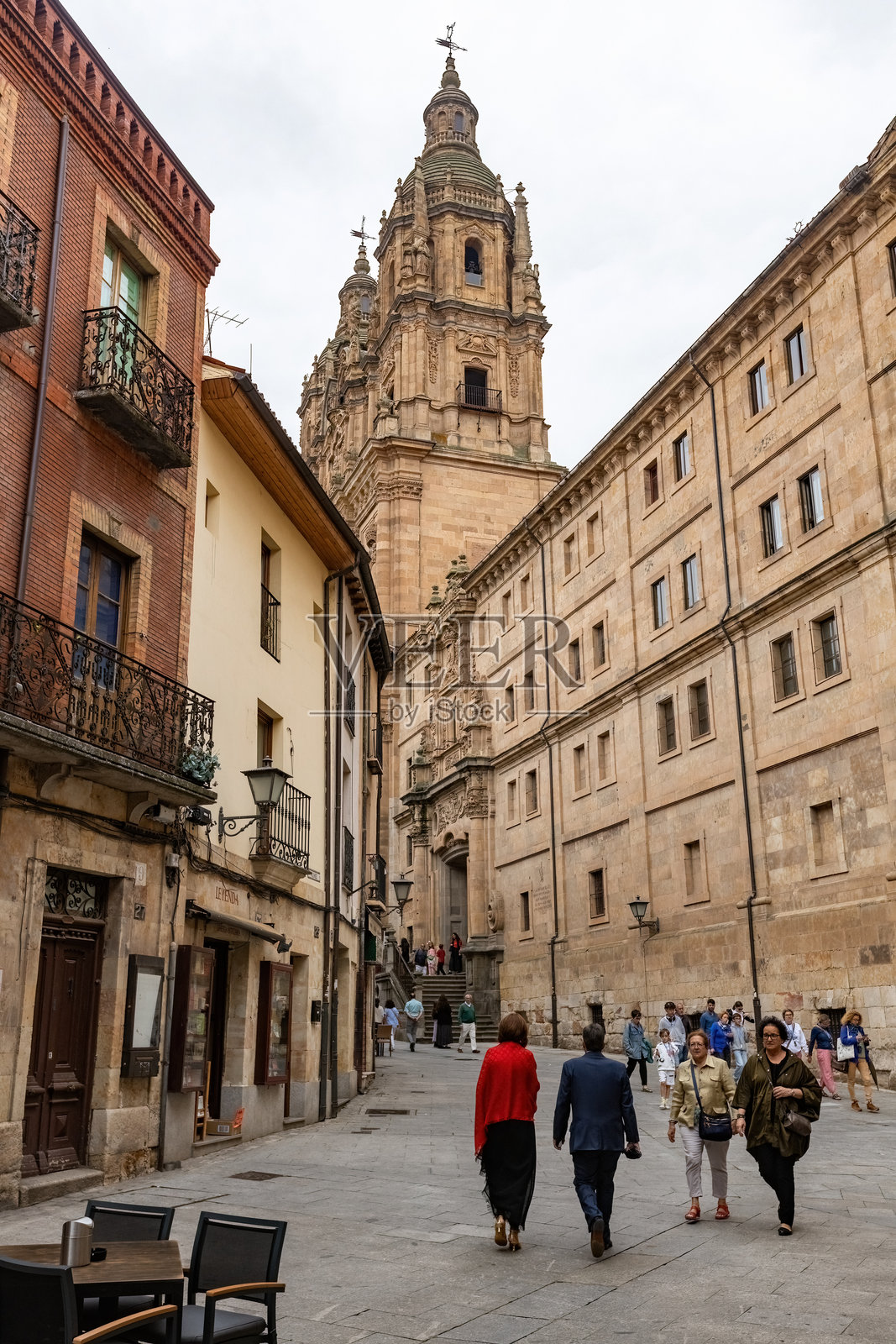 西班牙萨拉曼卡的老城区街道，人们在小镇上漫步，远处可见萨拉曼卡的拉克莱雷西亚天主教堂塔楼。照片摄影图片