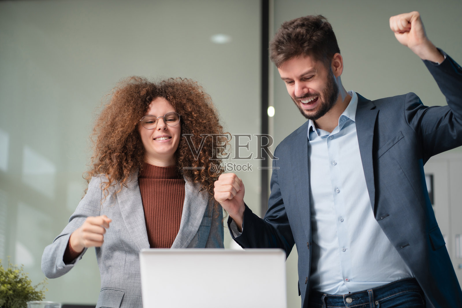 成功的商业团队，兴奋的年轻人们举起手臂，展现积极的情绪，在现代办公室里快乐地使用电脑工作。照片摄影图片