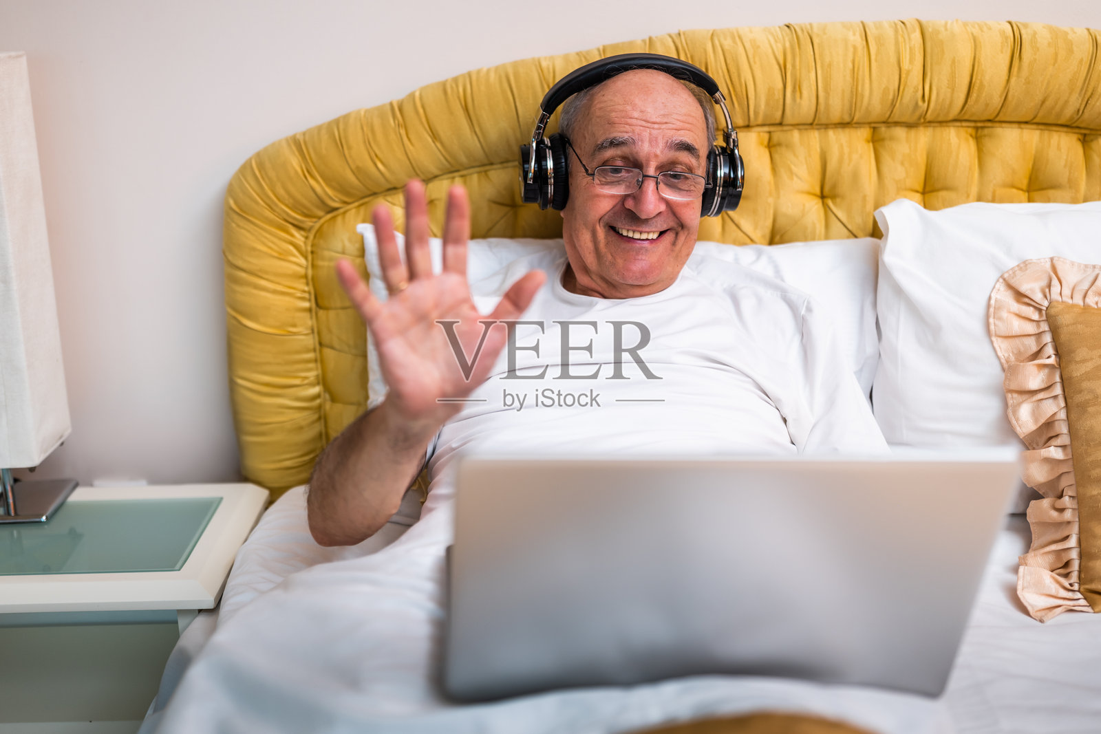 微笑的老人在床上通过笔记本电脑视频通话时戴着耳机并挥手照片摄影图片
