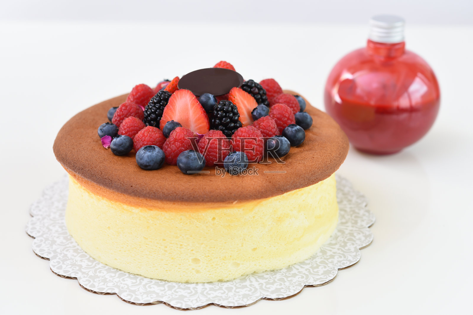 松软的日本芝士蛋糕，上面装饰着草莓、覆盆子、蓝莓和黑莓，旁边是一瓶红莓酱，特写侧视图展示了这款美味而受欢迎的甜点。照片摄影图片
