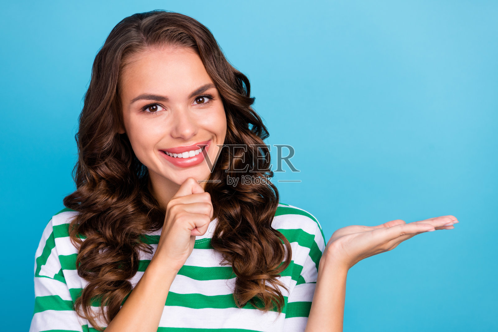 年轻的棕色卷发女士穿着条纹T恤，手持空白空间，背景为蓝色。照片摄影图片