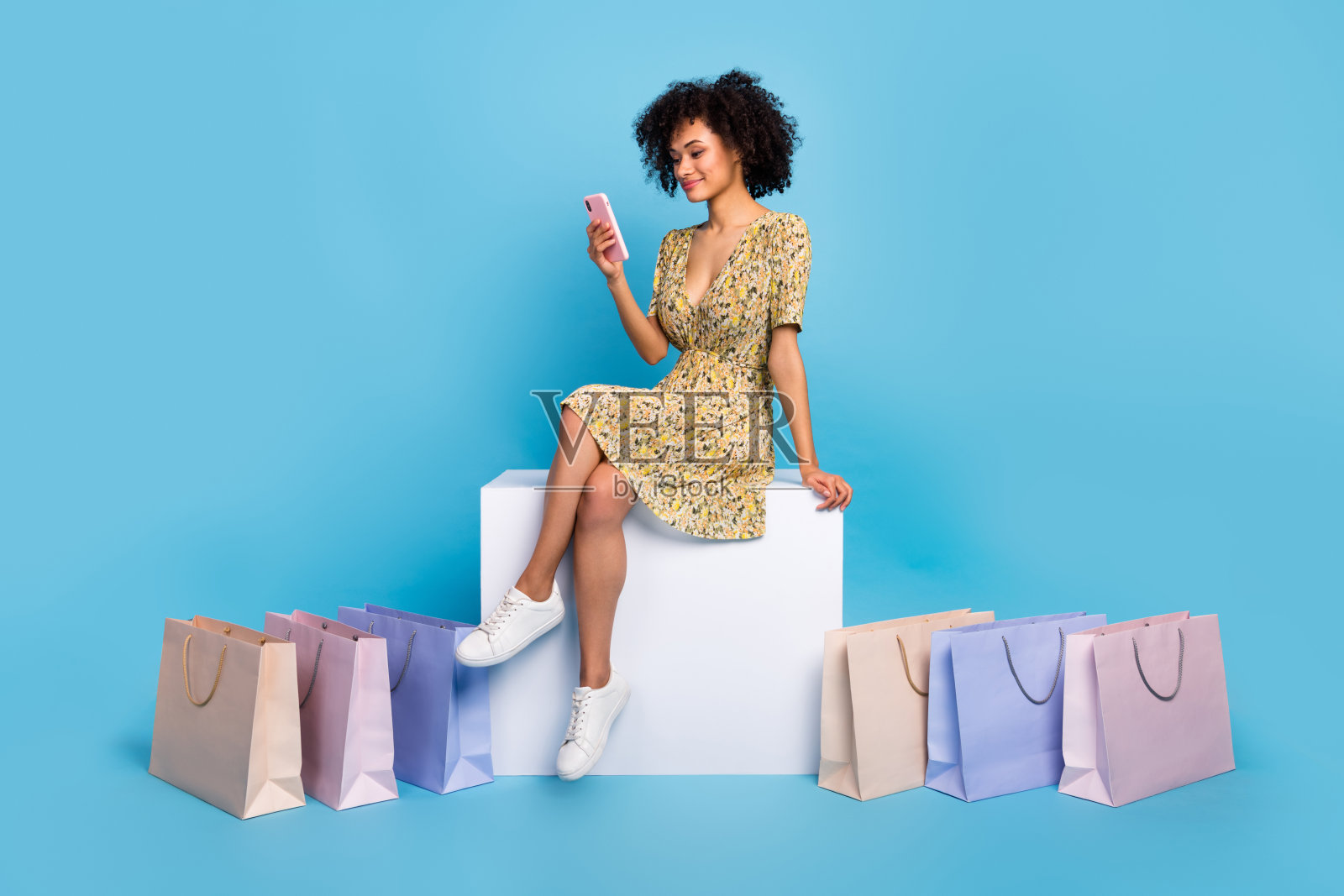 穿着印花服装的漂亮女士坐在方形椅子上，旁边有购物袋和现代设备，背景为蓝色。照片摄影图片