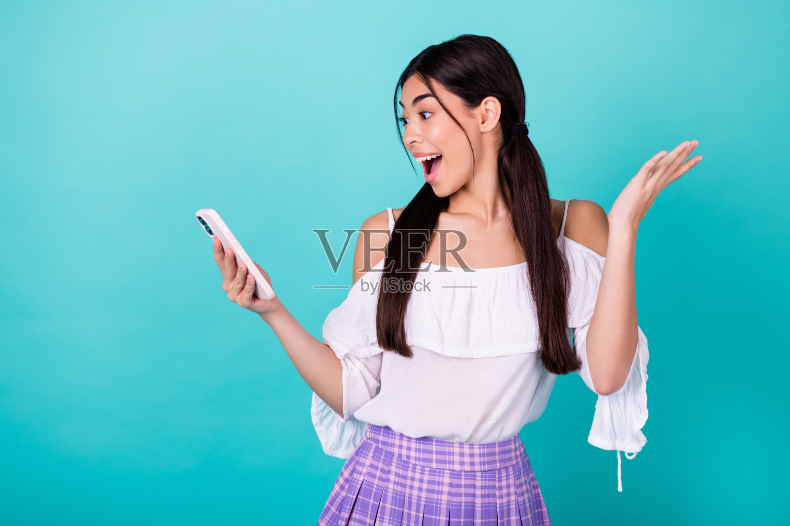 迷人兴奋的泰国女孩手持电话，惊讶地接收不可思议的消息，背景为青色。照片摄影图片