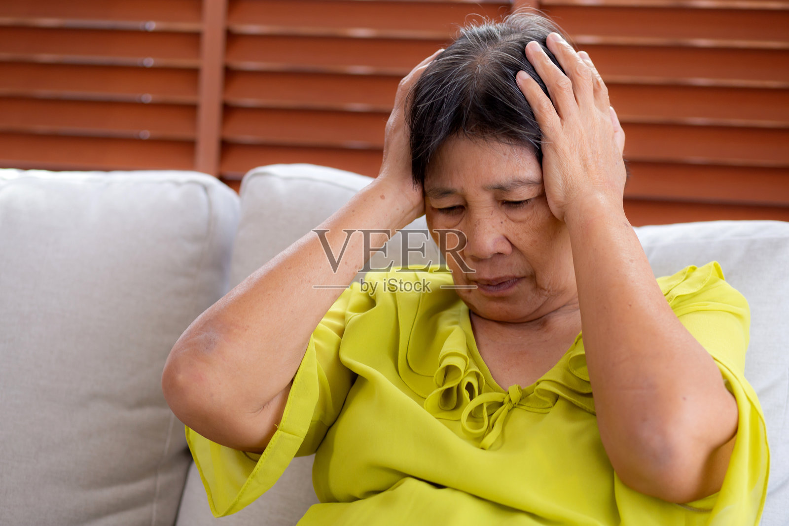 年长的亚洲女性坐在家中客厅的沙发上，感到疼痛和头痛，表现出抑郁和情绪压力，伴随偏头痛、沮丧和担忧，显得不快乐和疲惫。照片摄影图片