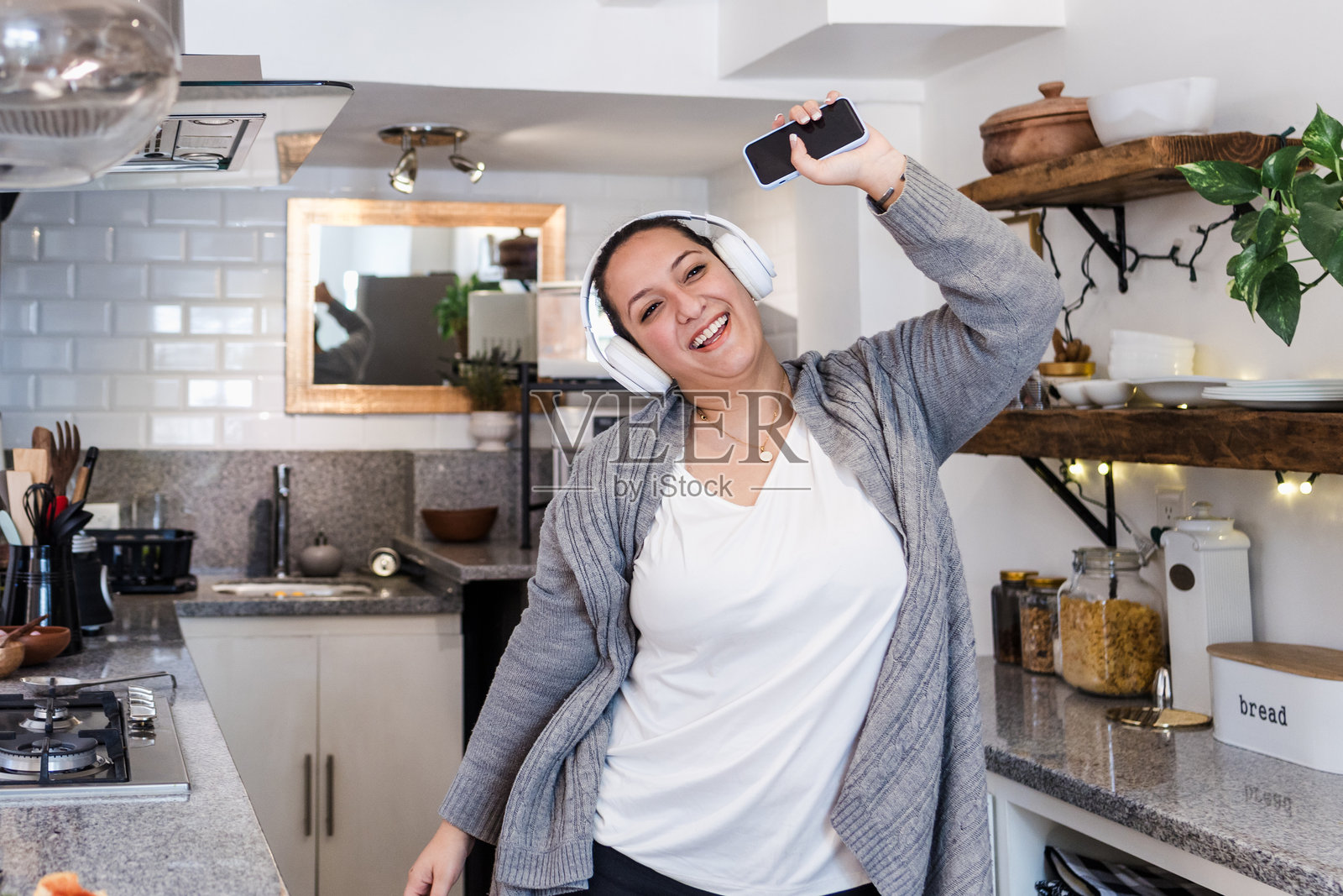 年轻的拉丁裔女性在墨西哥的家中厨房里，戴着耳机听音乐，手里拿着手机，跳舞并享受乐趣。照片摄影图片
