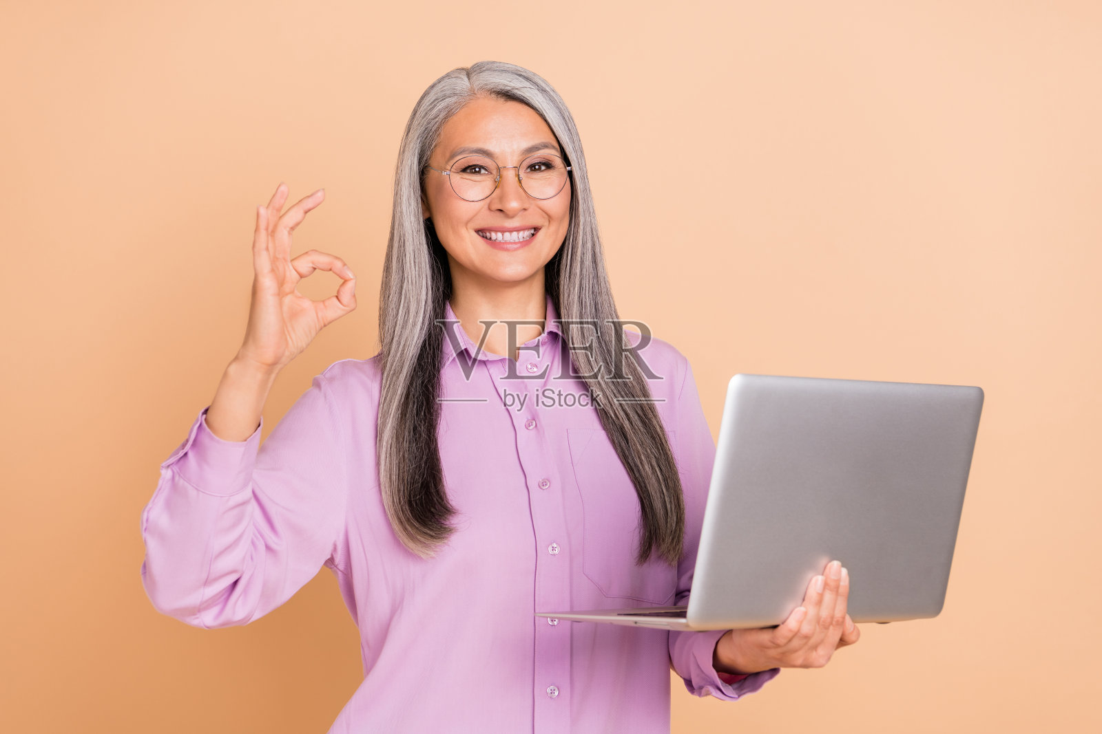一位酷炫灰发的女士戴着眼镜，穿着紫色衬衫，手持笔记本电脑，做出“好的”手势，背景为米色。照片摄影图片