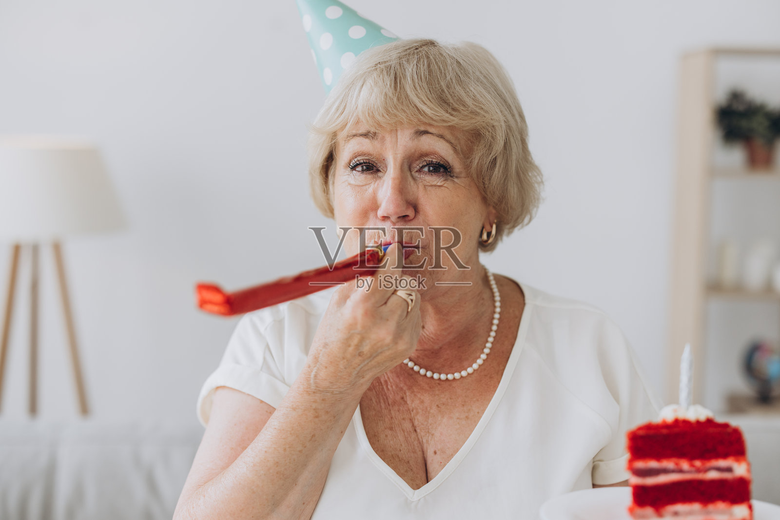 快乐的老年女性庆祝生日，戴着派对帽，手里拿着一个点着蜡烛的蛋糕，正在吹响喇叭。照片摄影图片