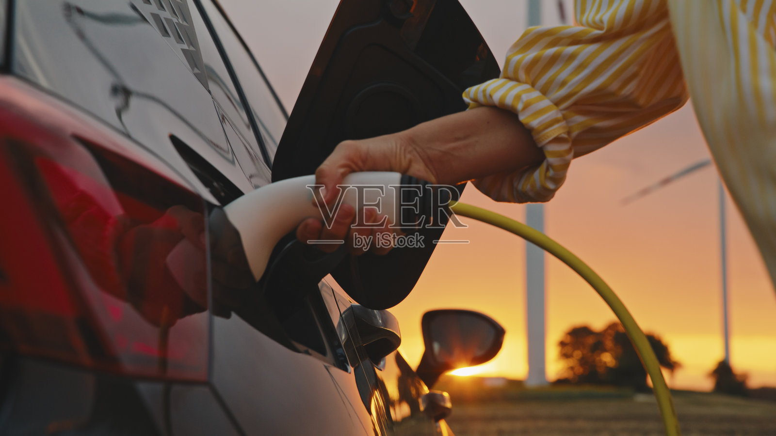 女性手在日落时分为电动车充电，背景是风力发电机。照片摄影图片