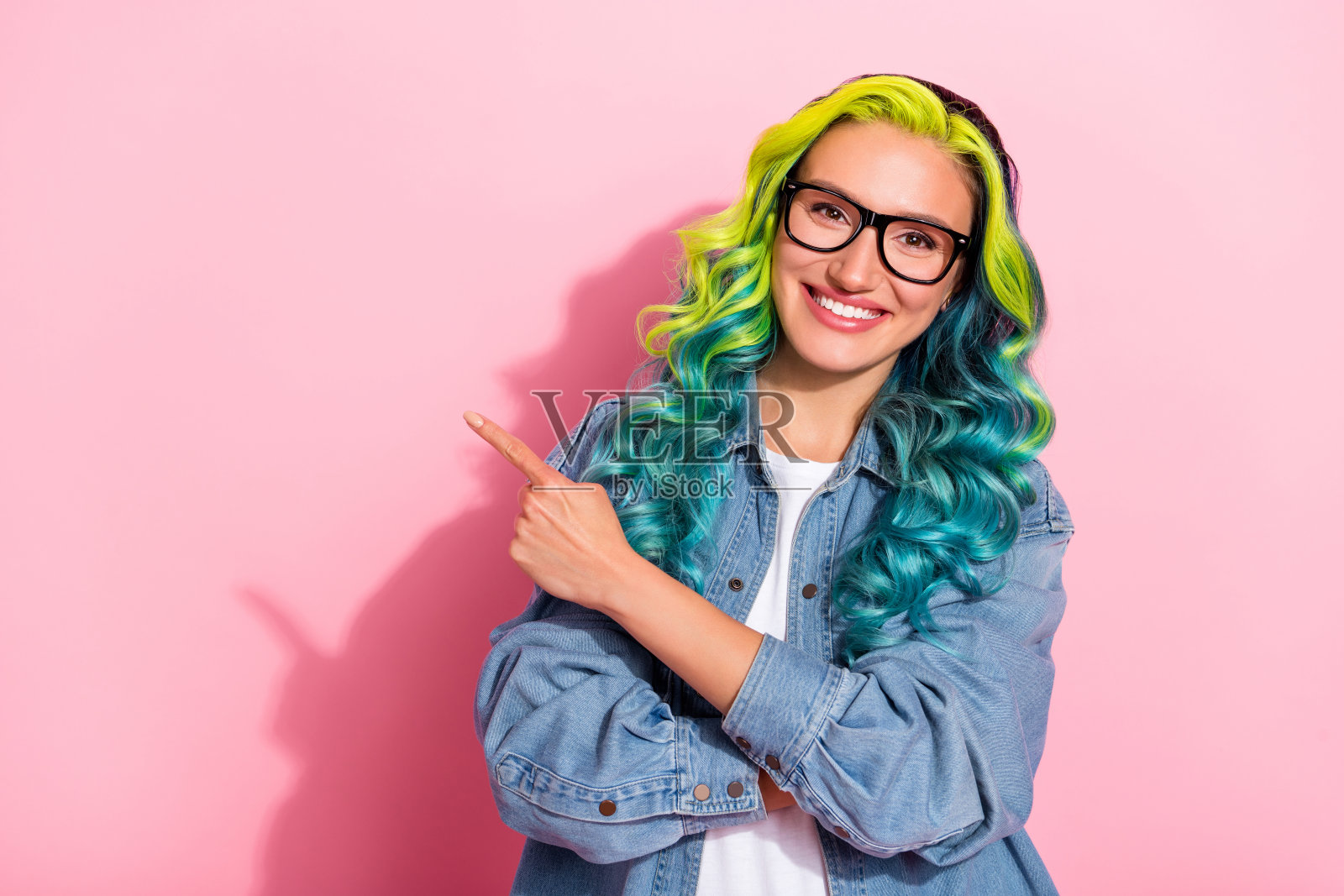 自信、光亮的年轻女士穿着牛仔夹克，戴着眼镜，微笑着指向粉色背景中的空白处。照片摄影图片