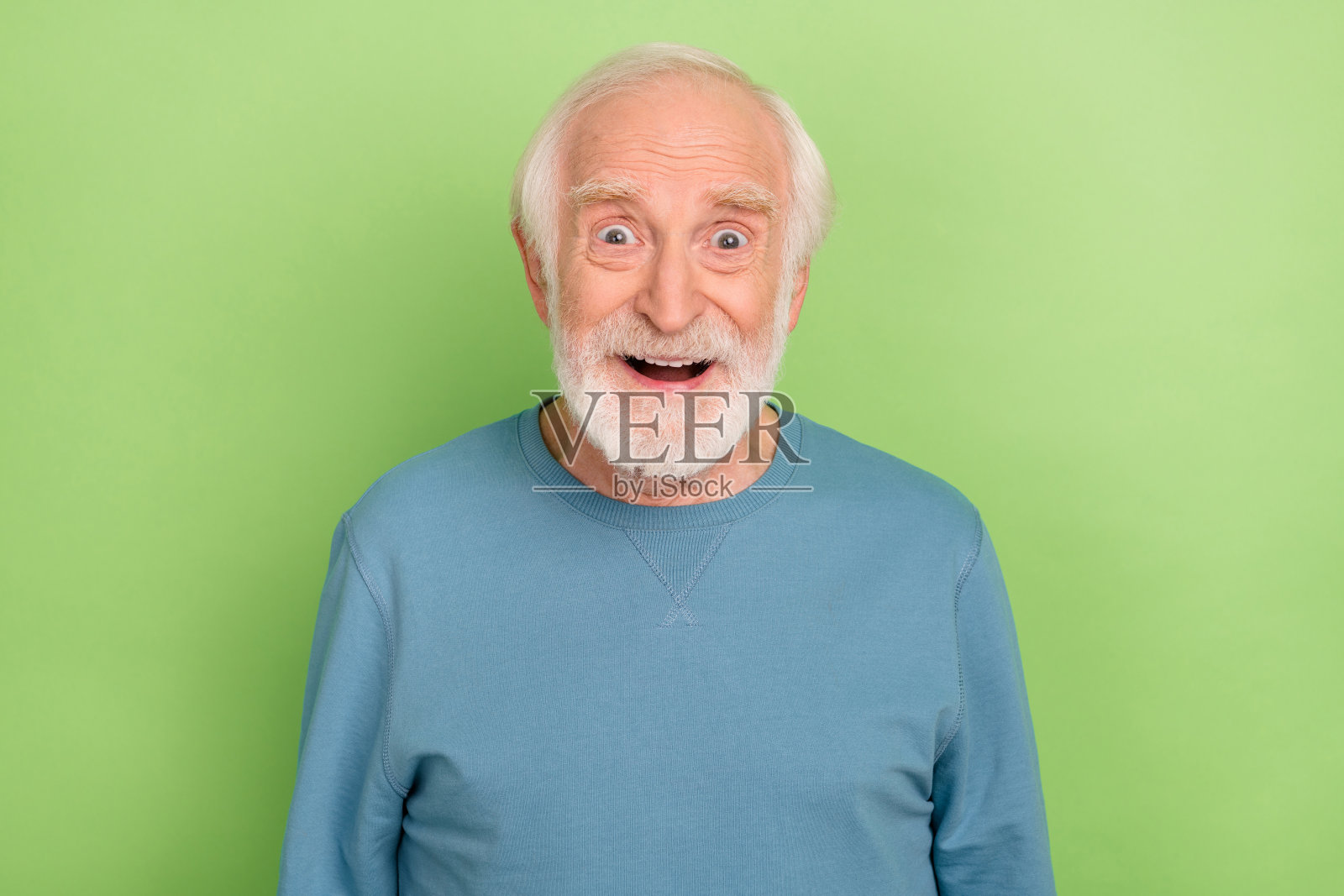 一位留着胡子的老者穿着蓝色毛衣，惊讶地看着镜头，背景为绿色。照片摄影图片