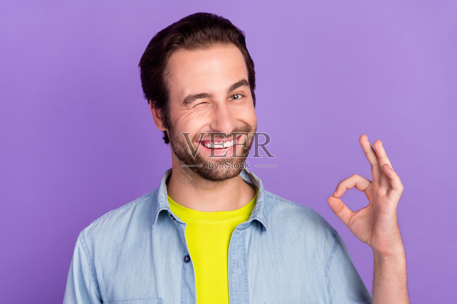 年轻帅气的男孩微笑着眨眼，调皮地做出“好的”手势，背景为紫色。照片摄影图片