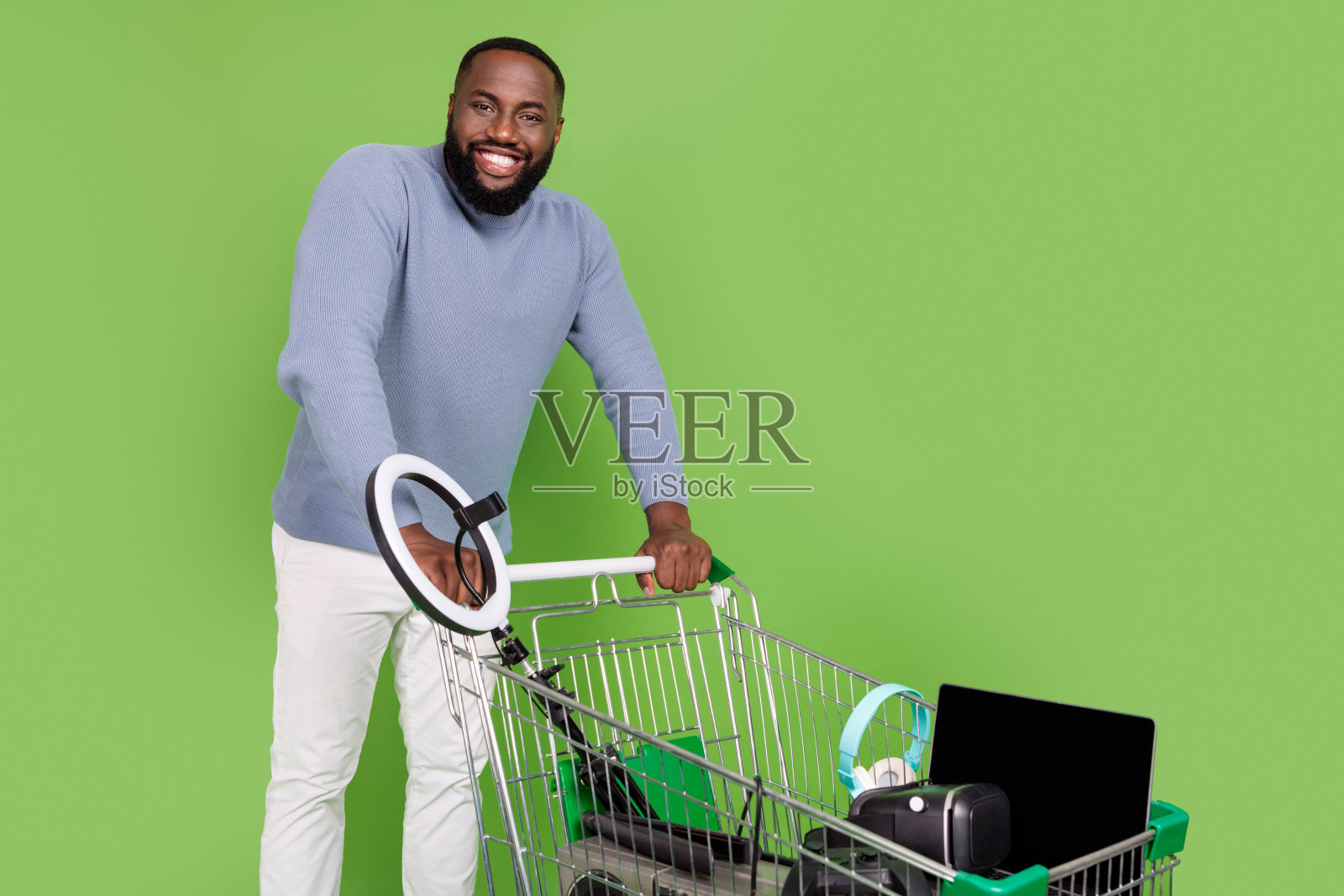 穿着毛衣的快乐非洲男士在绿色背景下走着，手里提着购物托盘，里面装满了各种小玩意儿。照片摄影图片