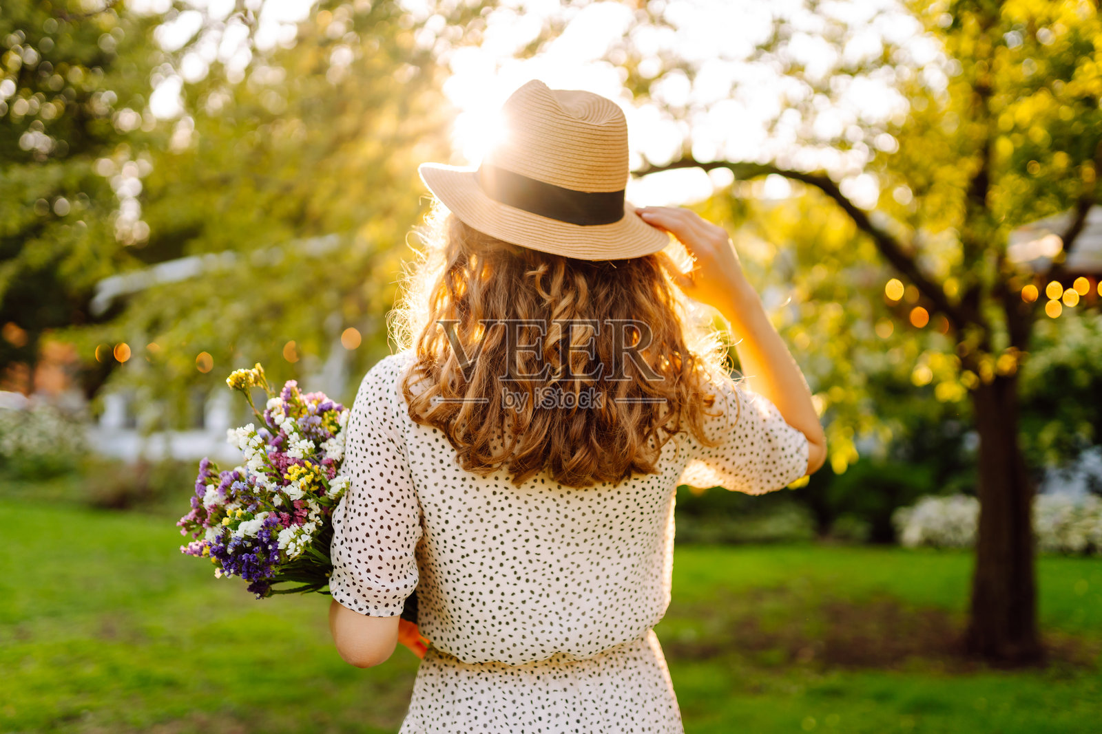 一位美丽的女性手持手机，手捧一束郁郁葱葱的野花，在绿色公园中享受日落。照片摄影图片