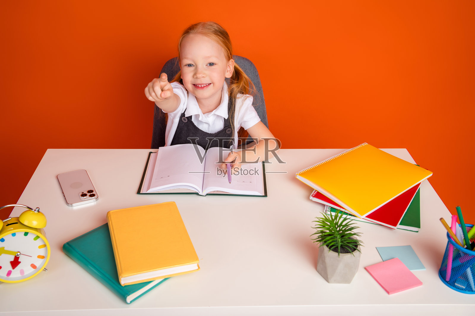 快乐的女学生在明亮的橙色背景下，兴奋地在桌子上学习，周围是五彩缤纷的学习用品。照片摄影图片