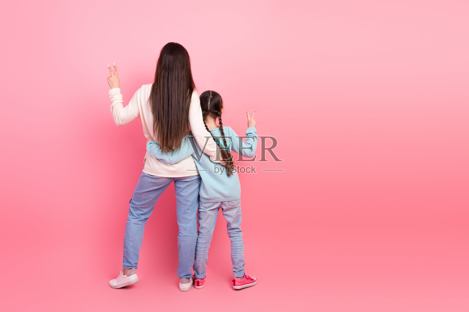 背影中，快乐的妈妈和女儿在鲜艳的粉色背景下比出和平手势，展现家庭乐趣。照片摄影图片