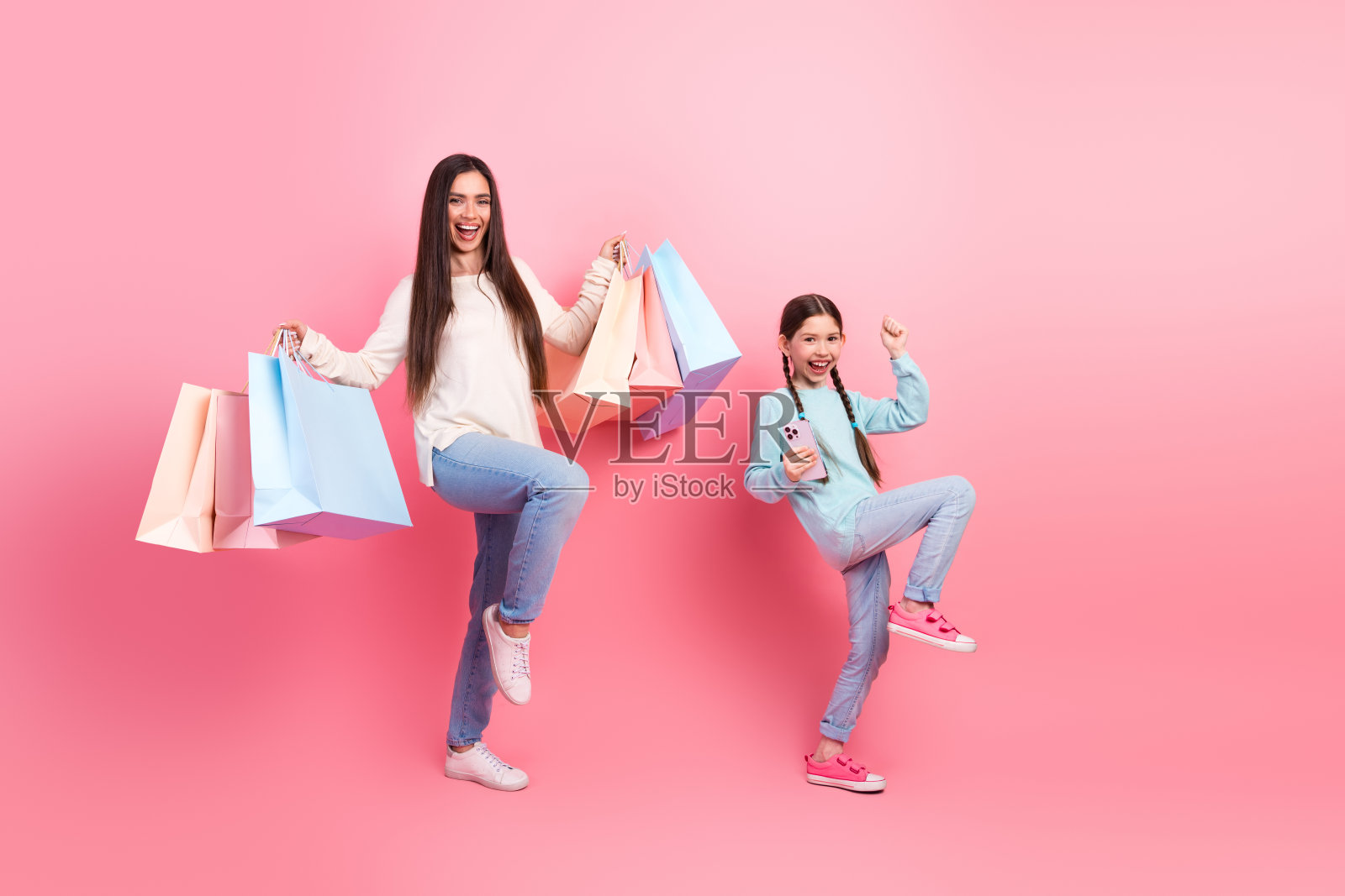 快乐的年轻母亲和女儿在充满活力的粉色背景下享受购物日，手里拿着购物袋和智能手机。照片摄影图片