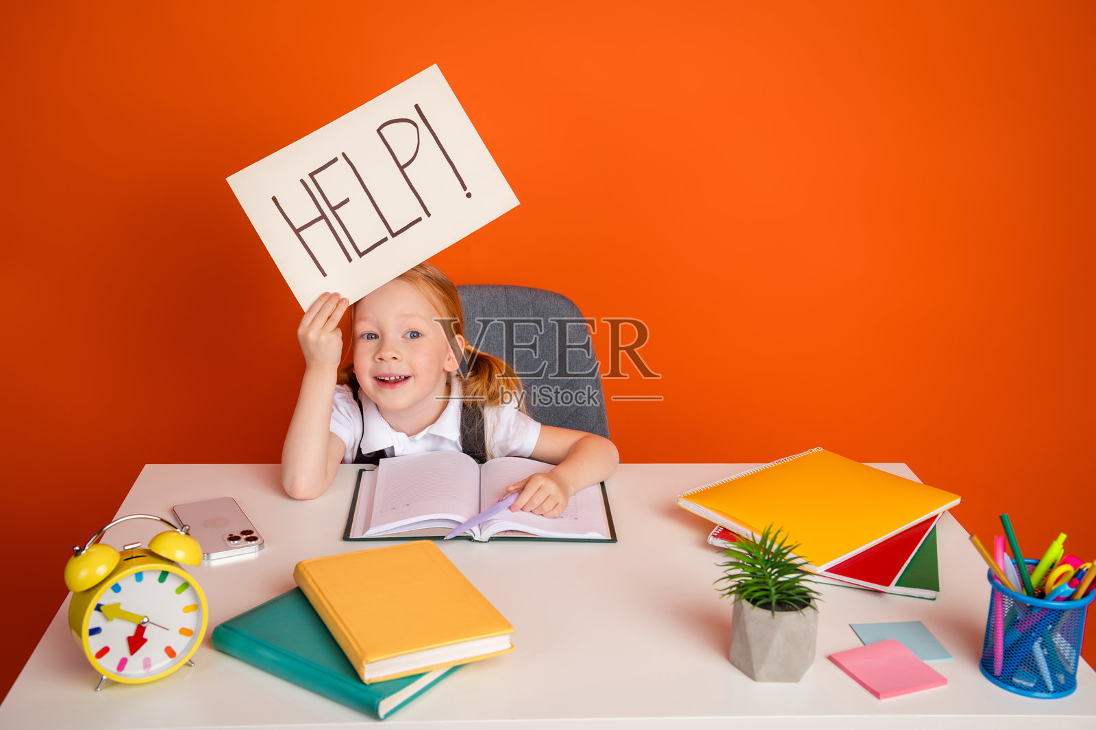快乐的女学生在书桌前举着求助标志，桌上有书籍和文具，背景为鲜艳的橙色。照片摄影图片