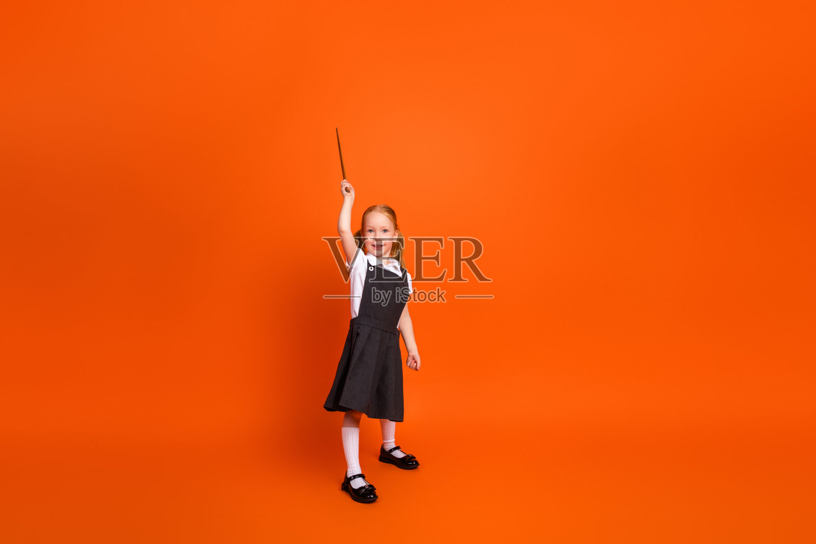 可爱的年轻女学生穿着校服，在鲜艳的橙色背景下举起魔杖，庆祝知识与学习。照片摄影图片