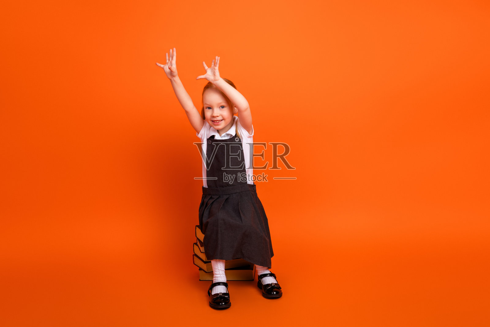 穿着校服的迷人年轻女学生坐在书本上，背景为橙色，面带愉快的微笑，双手举起。照片摄影图片