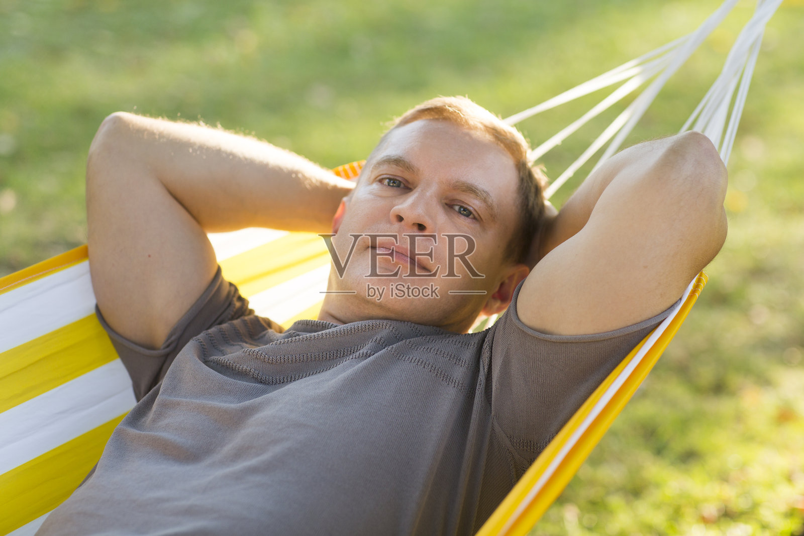 成熟微笑的男人躺在花园的秋千椅上，夏日阳光下注视着镜头。照片摄影图片