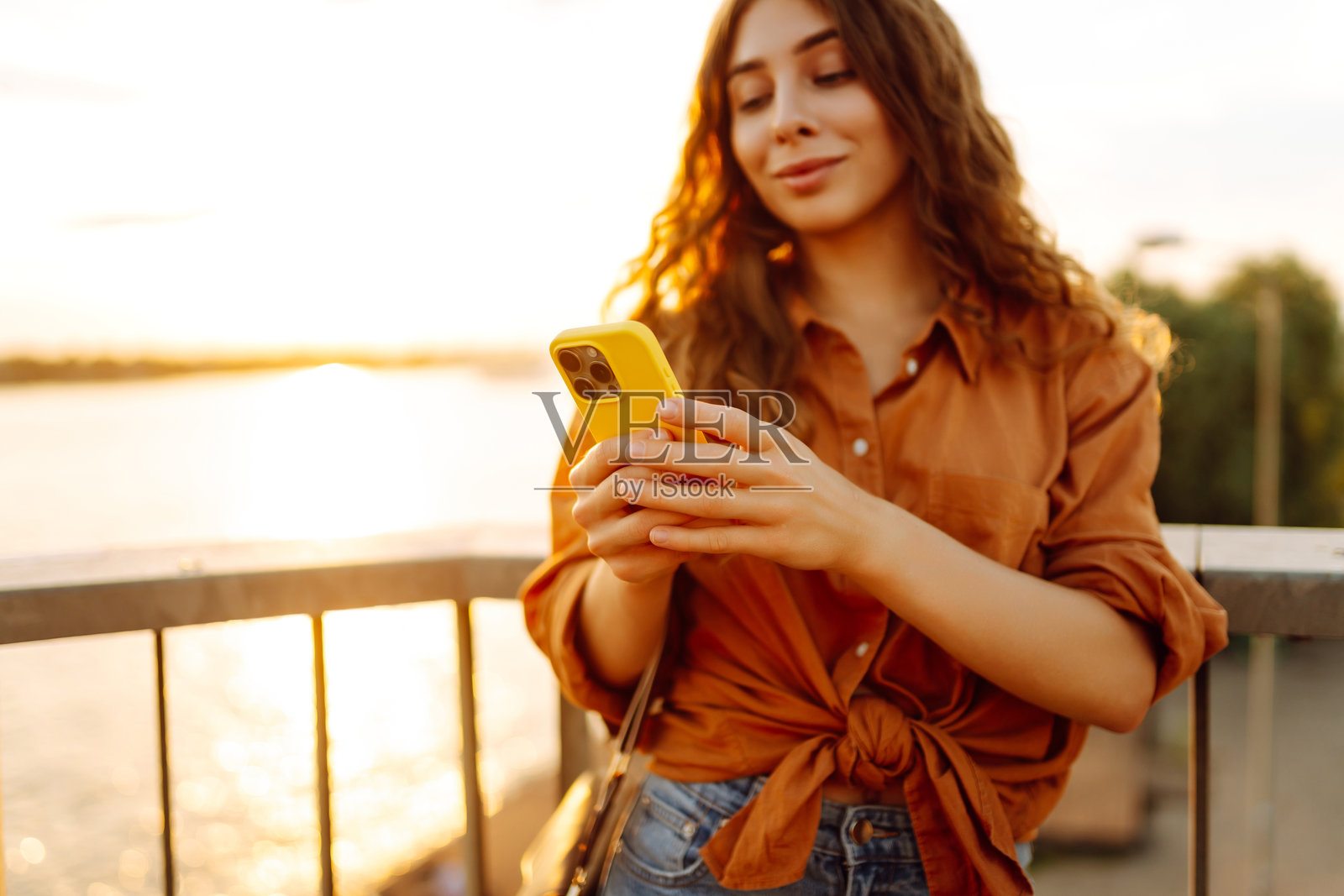 一位年轻女性手持手机，走在日落时分的堤岸上。照片摄影图片