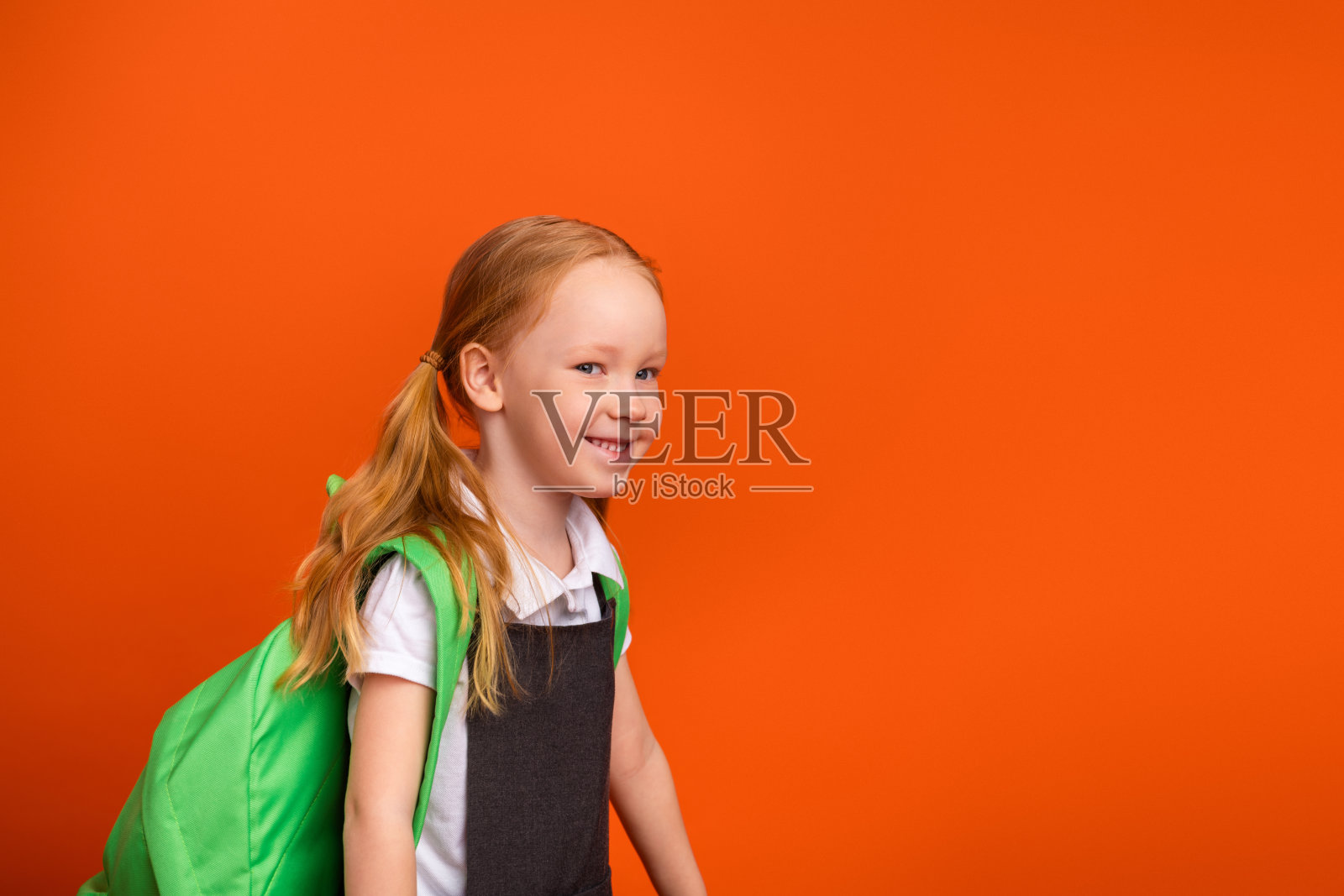 可爱的红发女学生背着绿色书包，在橙色背景前微笑，摆出愉快的姿势照片摄影图片