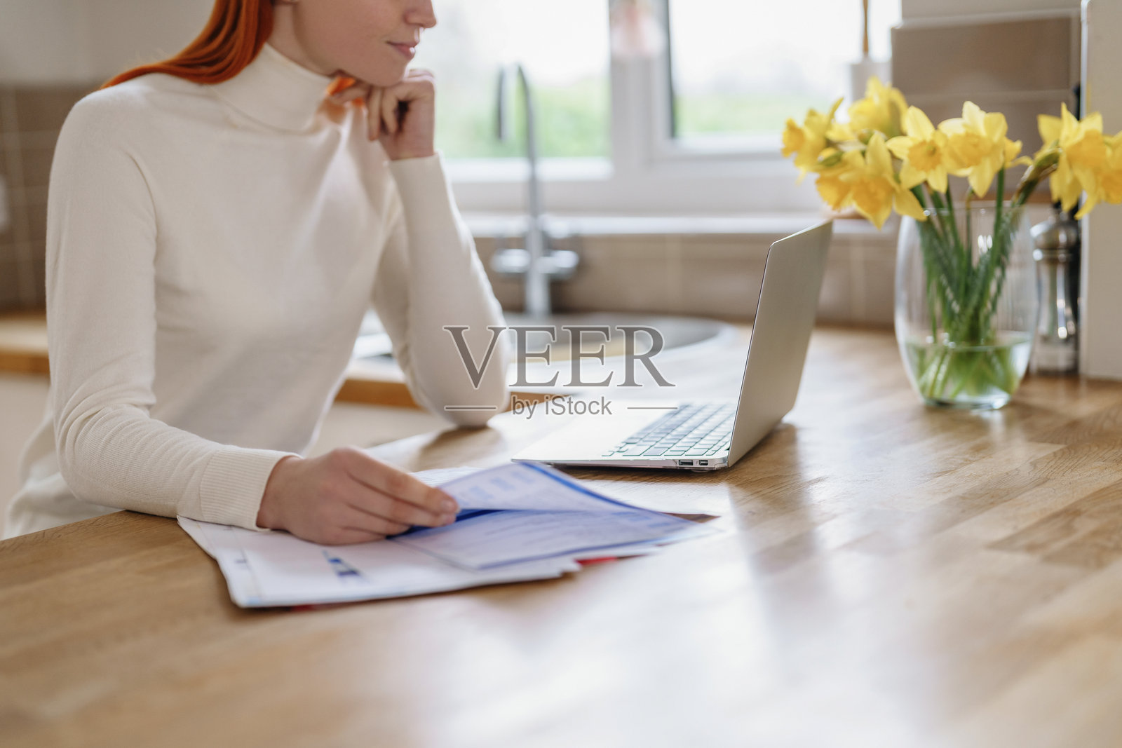 年轻女性在明亮的厨房中使用笔记本电脑审阅文件，桌上摆放着花朵，白天的光线洒入室内。照片摄影图片