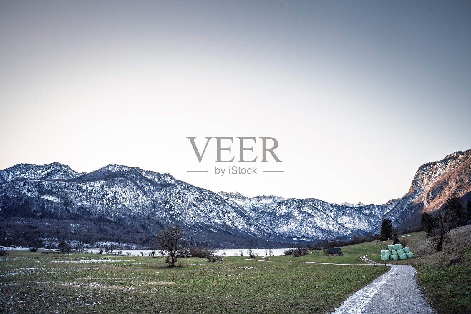 斯洛文尼亚博欣湖的初冬全景，雪覆盖的朱利安阿尔卑斯山矗立在绿色农田之上，干草卷和一条通往宁静湖水的农场小路在清澈的柔和天空下延伸。照片摄影图片