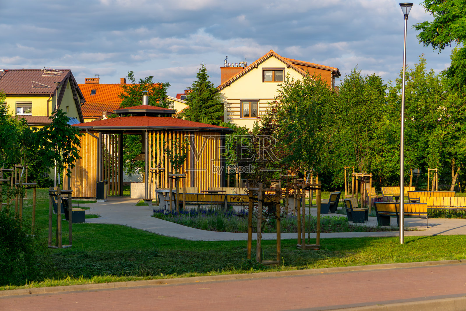 现代木制凉亭位于城市公园中，周围有年轻的树木、长椅和步道。背景中可见独栋住宅和路灯。照片摄影图片