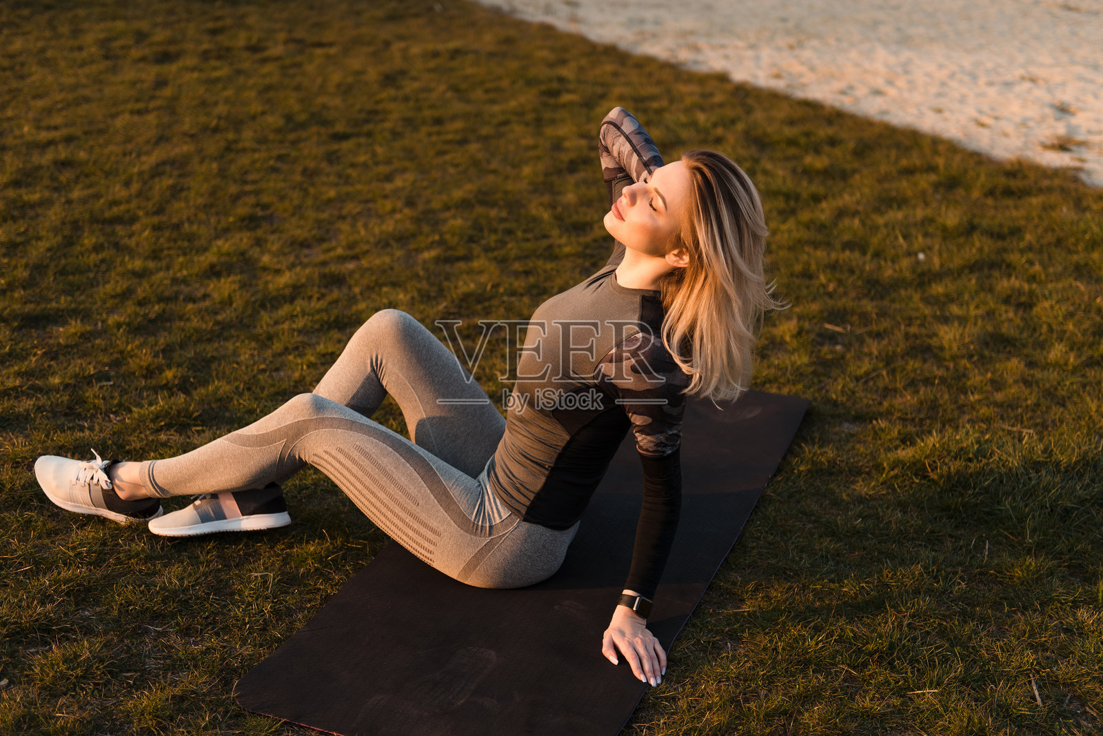运动年轻女性坐在游乐场附近的瑜伽垫上，闭着眼睛，手中拿着手机。照片摄影图片