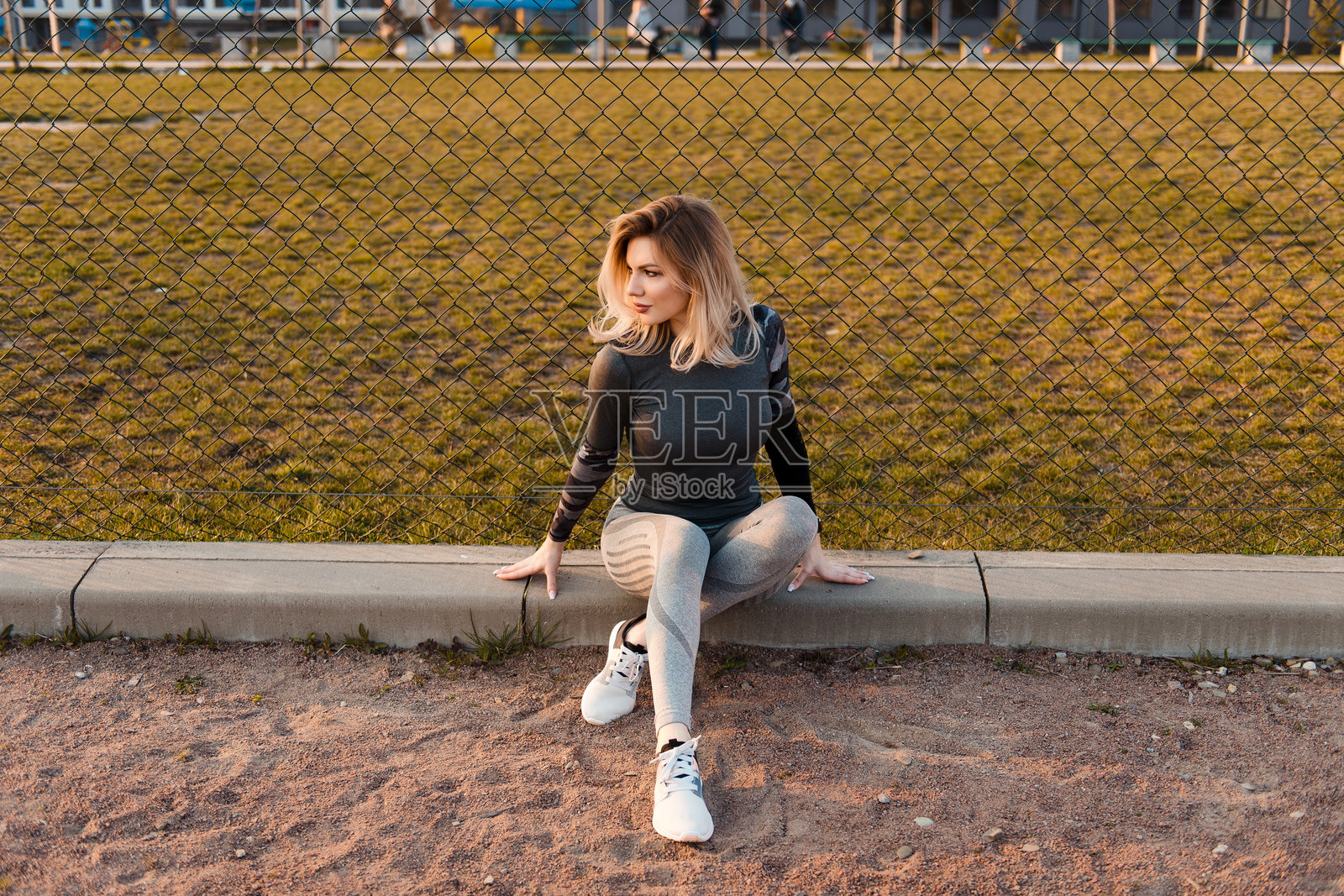 运动型金发女性坐在人行道边，靠近铁丝网，目光投向远方。照片摄影图片