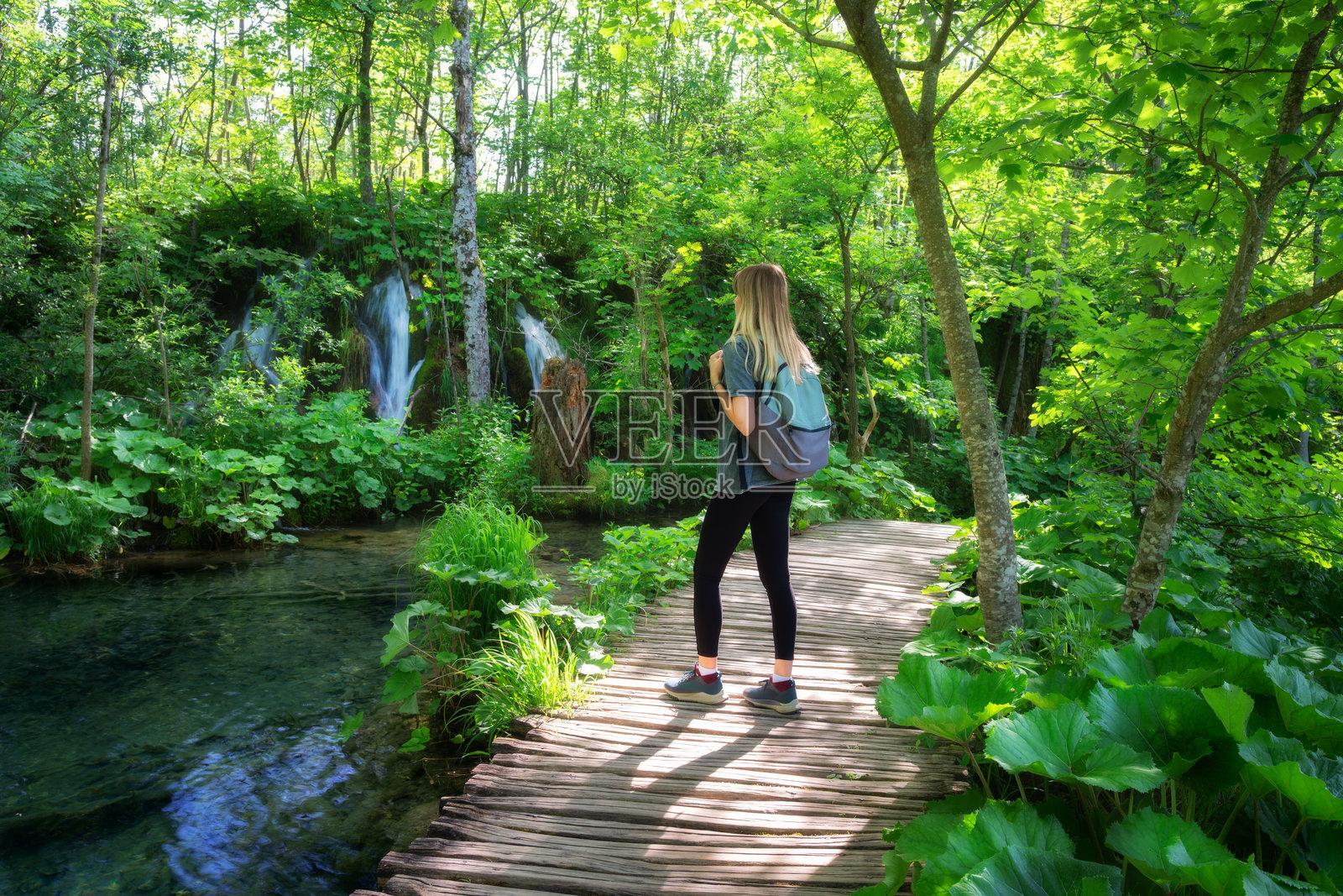 背着背包的年轻女性在克罗地亚普利特维采湖国家公园的木栈道上行走，身边是壮观的瀑布和绿色的树木，夏季的日落时分。女游客。运动女孩在森林中徒步旅行。照片摄影图片