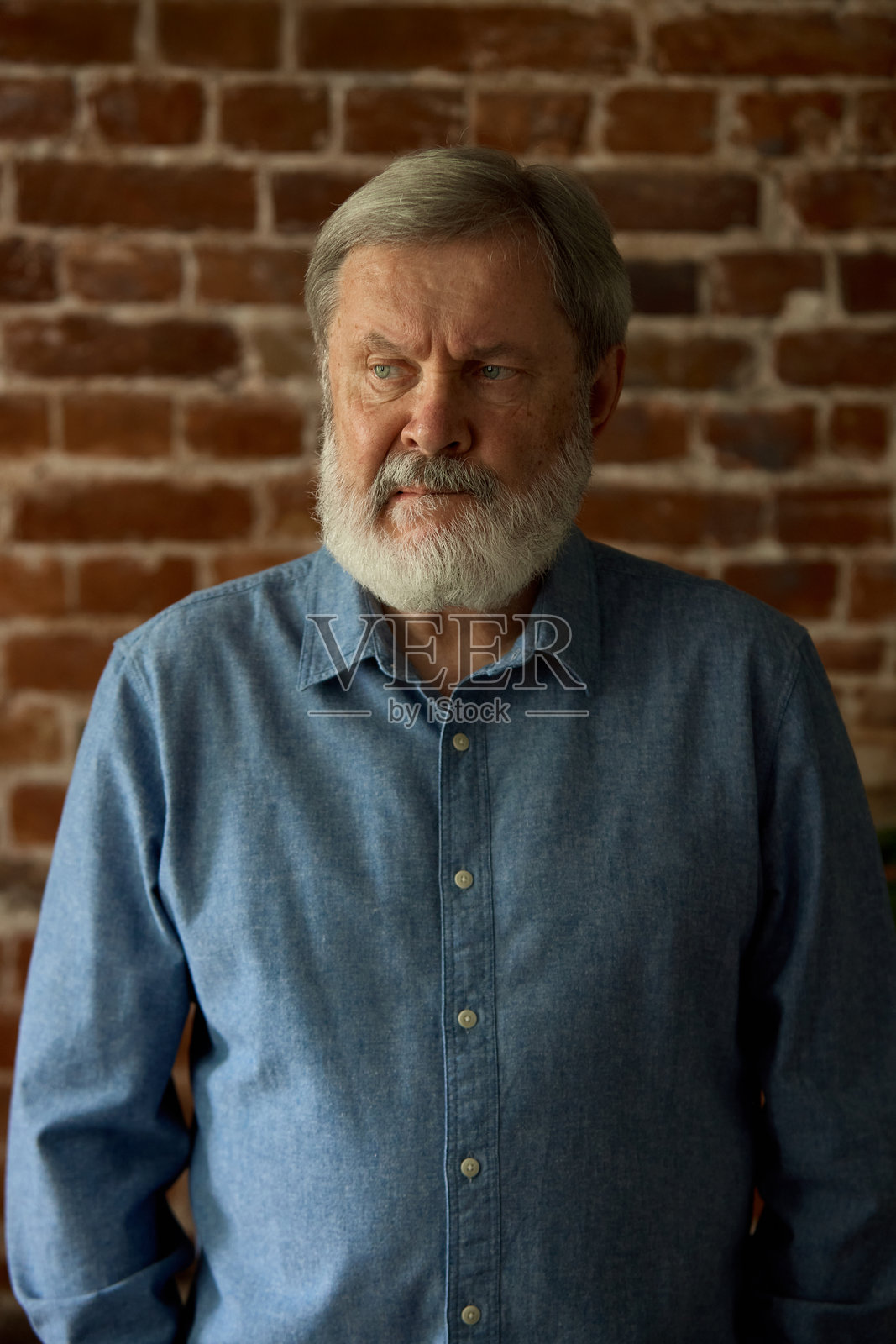 身穿牛仔衬衫的老年男性侧身站立，面带平静自信的表情，背景是砖墙。稳重的气质。照片摄影图片