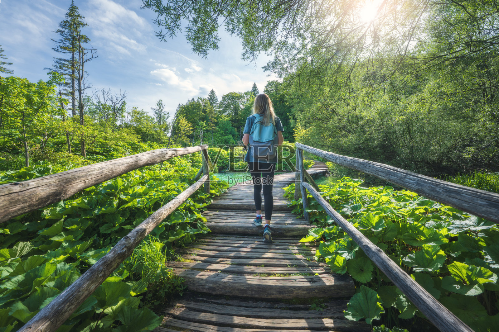 背着背包的年轻女性在克罗地亚普利特维采湖国家公园的木栈道上行走，身边是壮观的瀑布和绿色的树木，夏季的日落时分。女游客。运动女孩在森林中徒步旅行。照片摄影图片