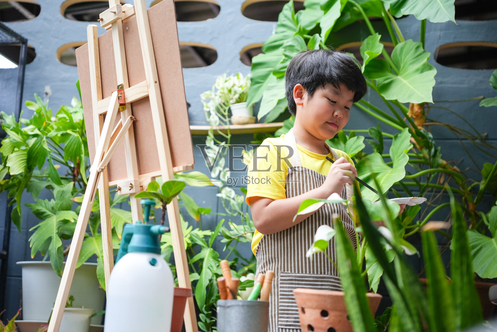 年轻的亚洲男孩在植物间户外绘画，创意儿童在绿色花园中使用画架和画笔，小艺术家在郁郁葱葱的户外环境中享受绘画。照片摄影图片