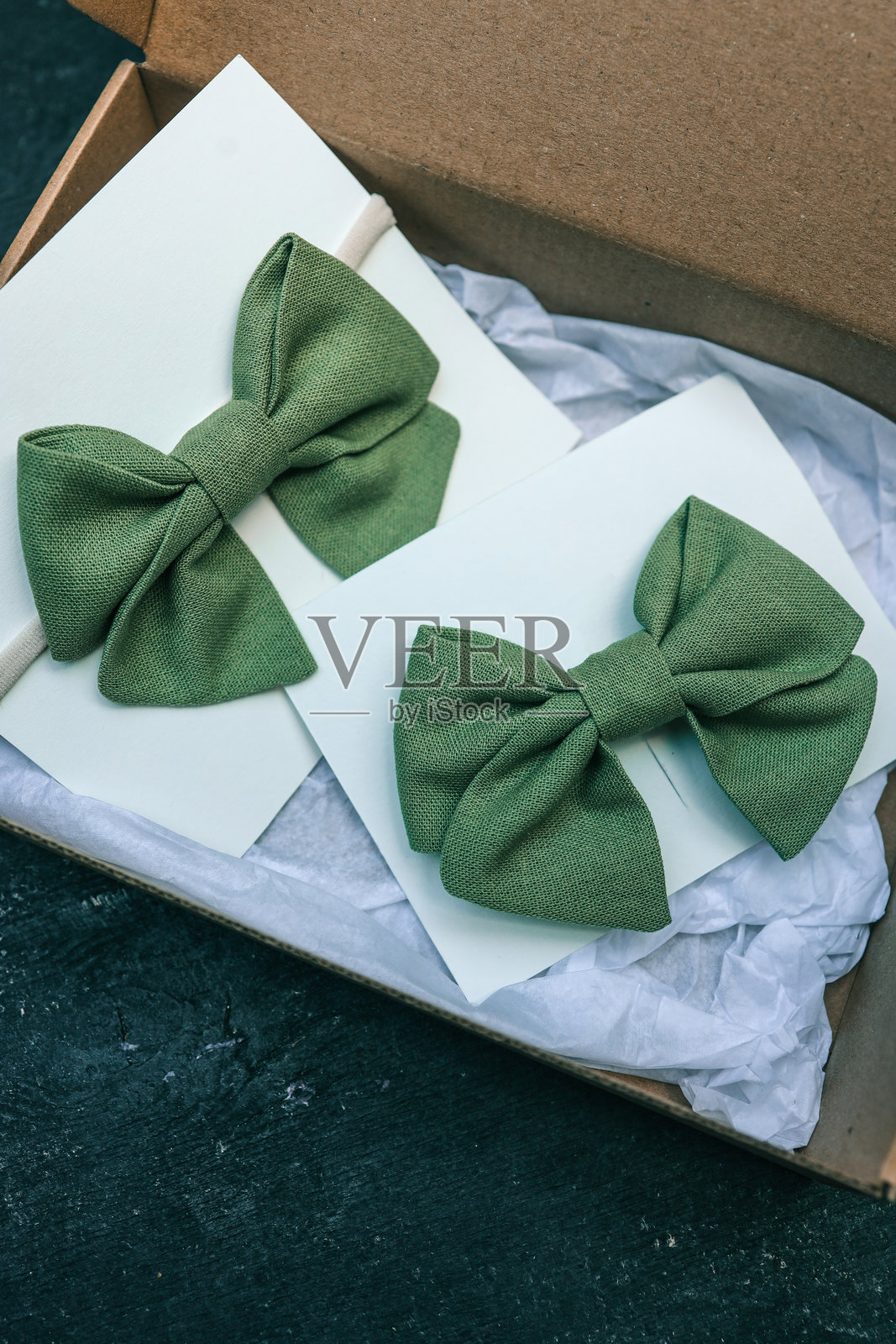 两条绿色亚麻蝴蝶结领带展示在白色卡片上，一条带有松紧带，另一条用夹子固定，风格为礼品盒。适合儿童或特殊场合的优雅手工配饰，色调柔和自然。照片摄影图片