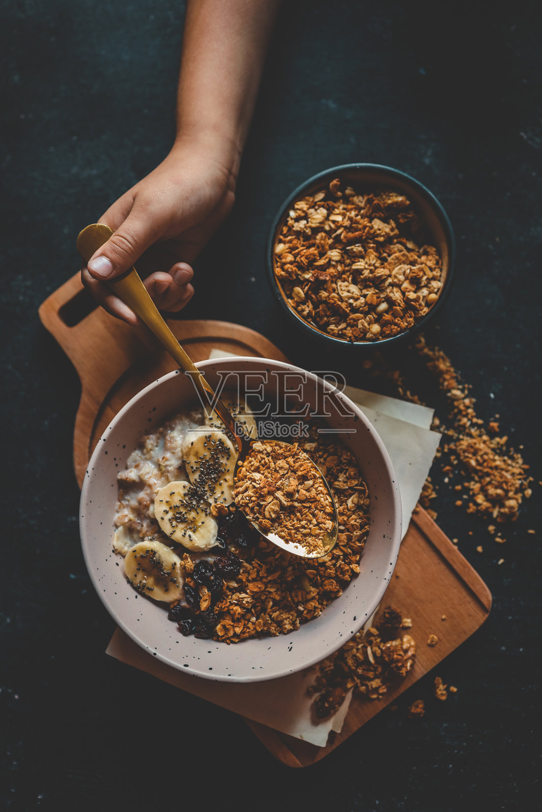 孩子的手握着一把勺子，勺子里装着格兰诺拉麦片，正对着一碗上面放有香蕉片和干果的燕麦粥，背景为深色。健康早餐概念，配有戏剧性的灯光。照片摄影图片
