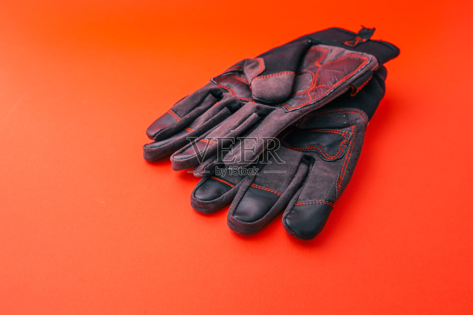 黑色和灰色带红色缝线的加固掌心和手指的防护工作手套，放置在鲜艳的红色背景上，适用于工业或建筑安全。照片摄影图片